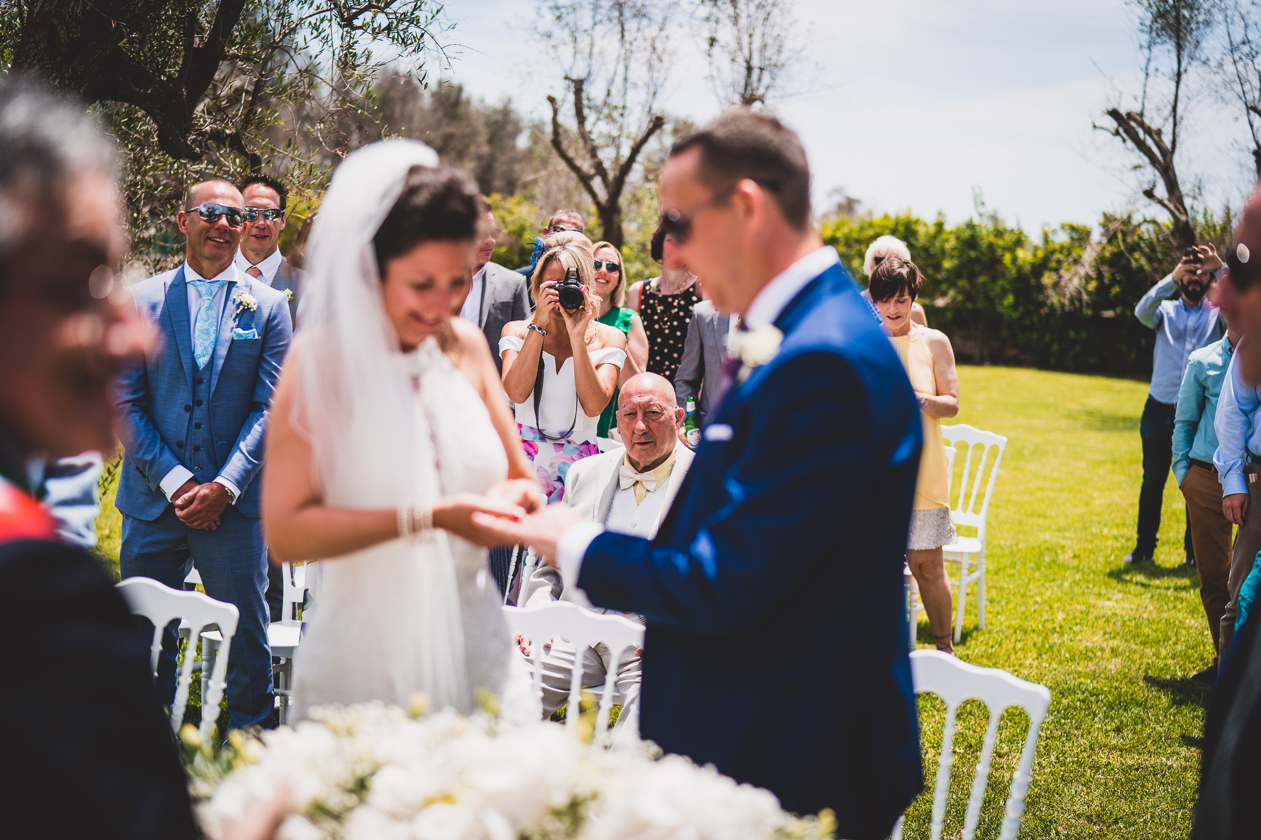 A wedding photographer captures the moment a bride and groom exchange their wedding rings in an outdoor ceremony.