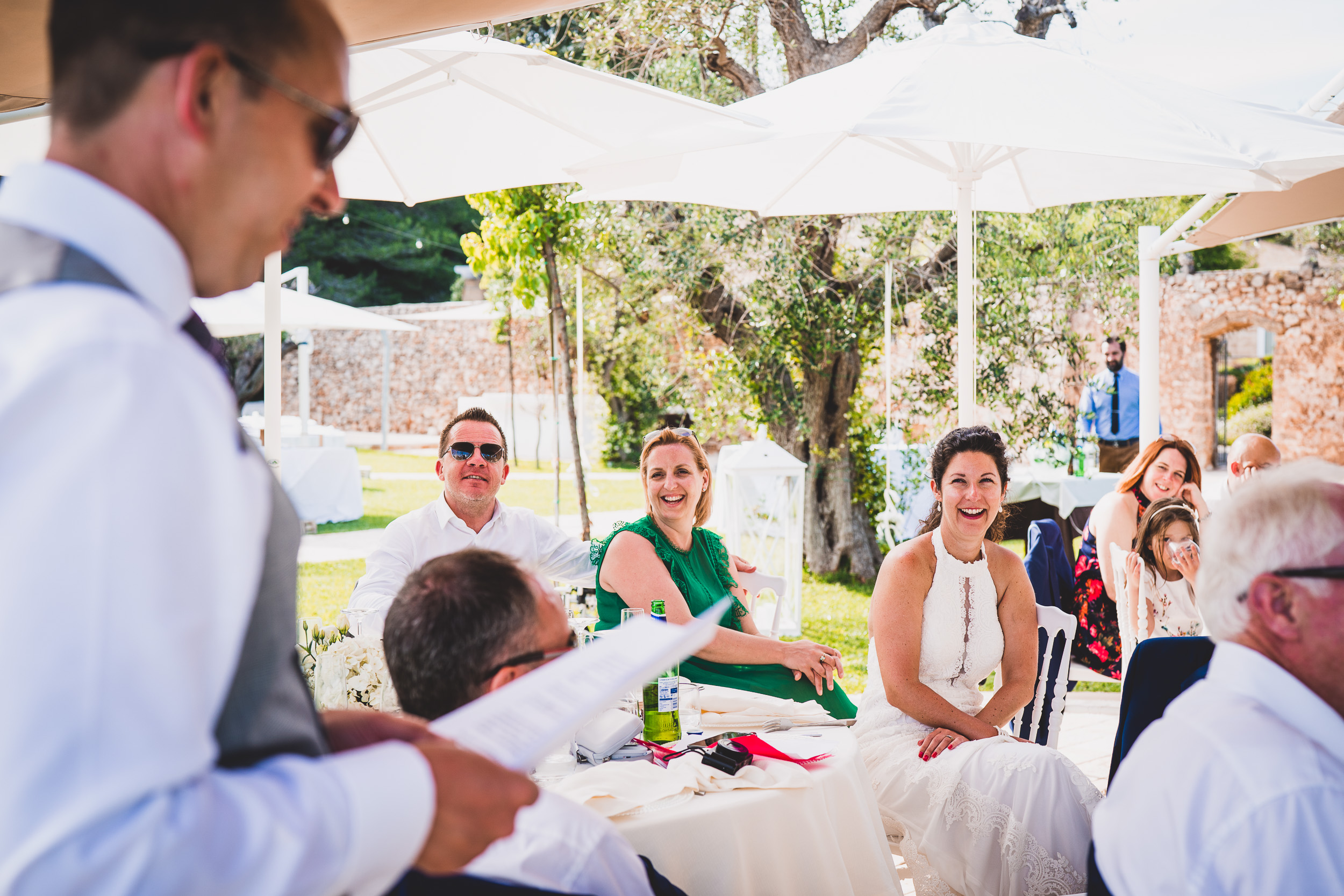 A man is speaking to a group of people at a wedding while referencing the bride and wedding photo memories.