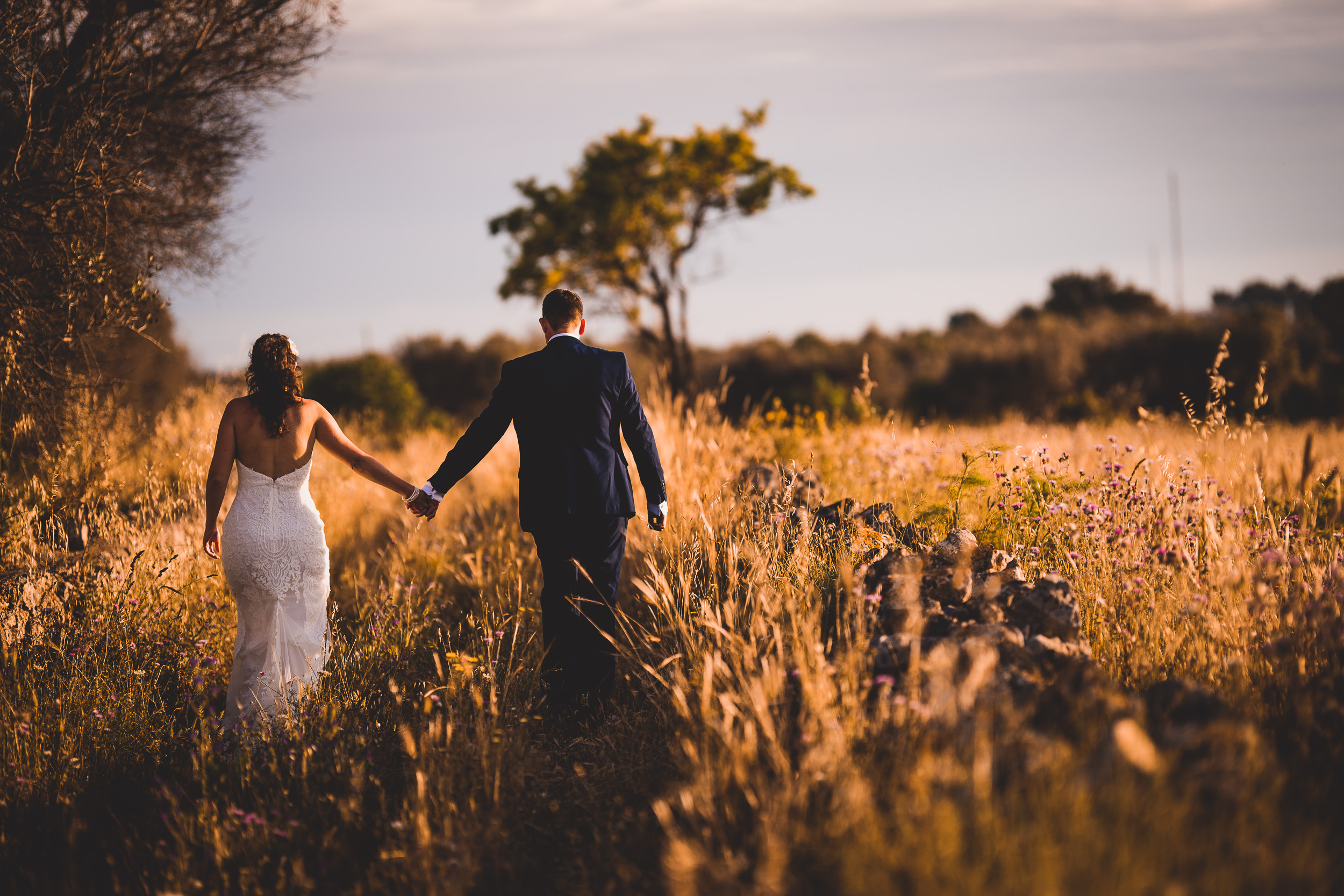 A bride and groom, captured by a wedding photographer, walking through a field of tall grass.