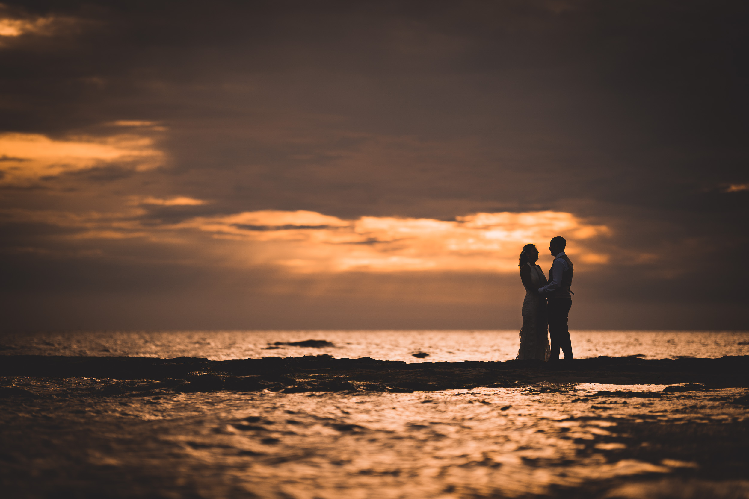 A wedding couple poses on a rock during sunset while their wedding photographer captures the moment.