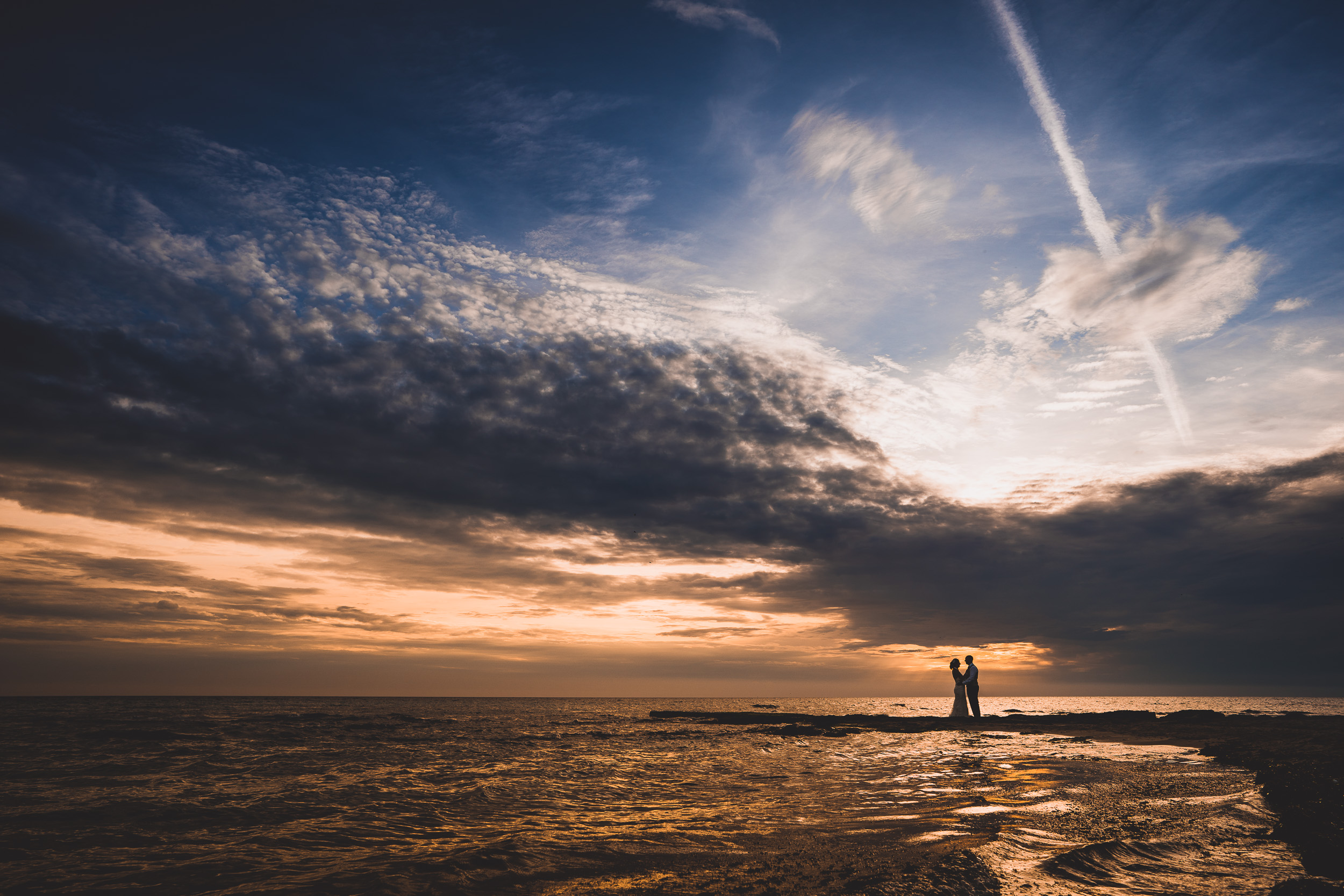 A bride and groom posing for a wedding photographer on the beach at sunset.