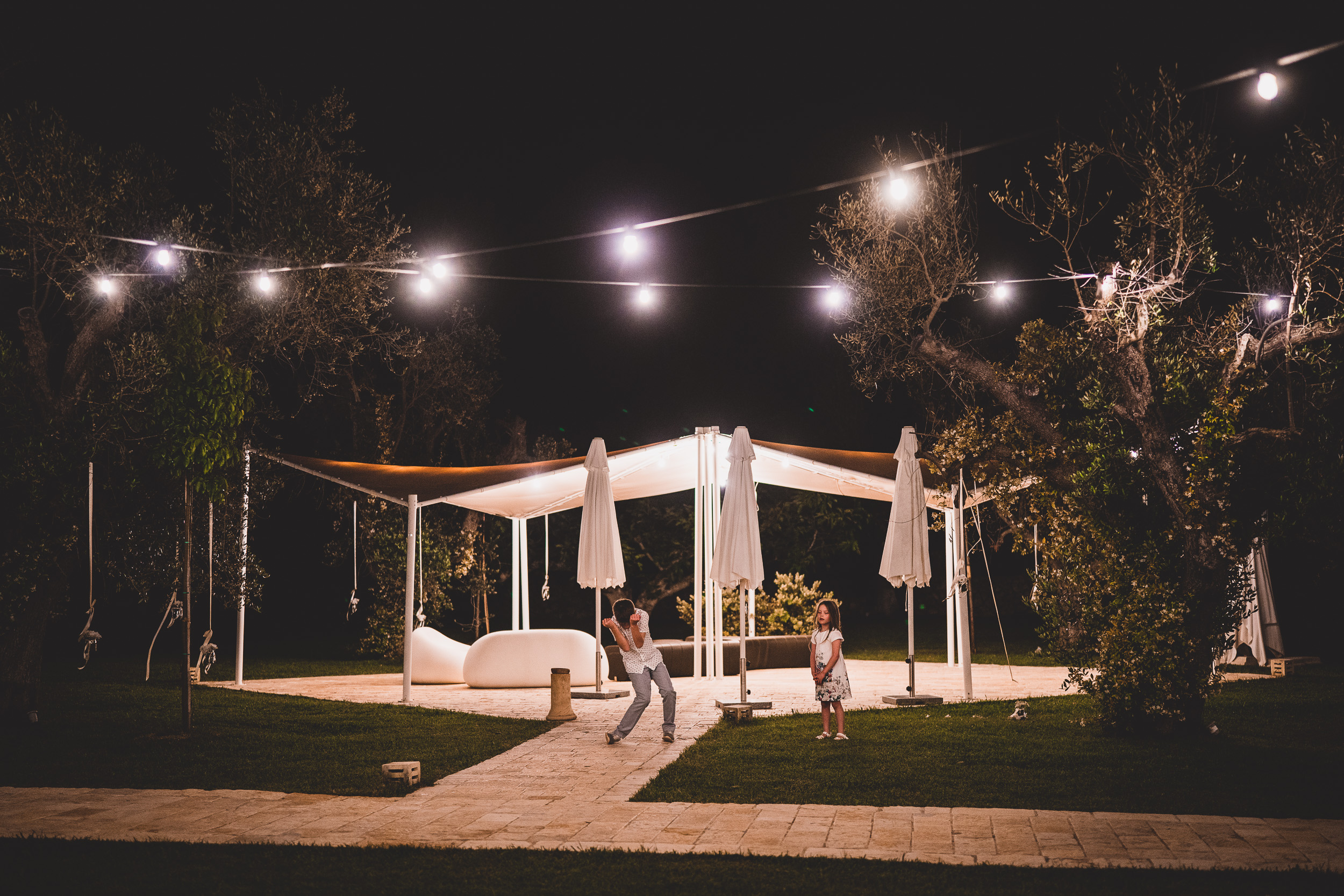 A bride stands under a string of lights in a garden.