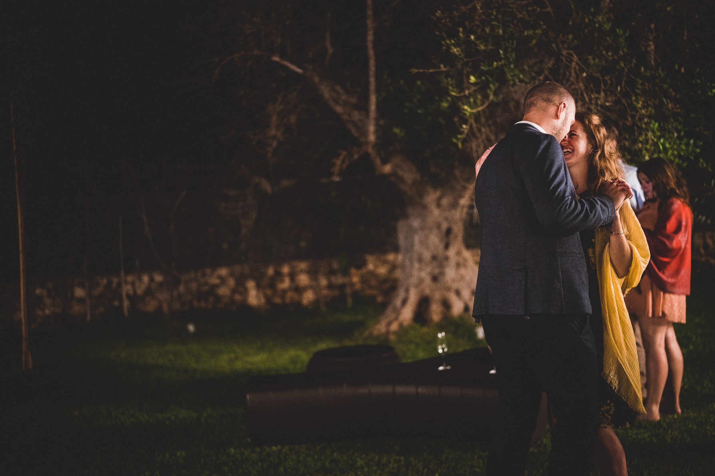 A wedding photographer captures a bride and groom kissing under a tree during their night-time wedding.