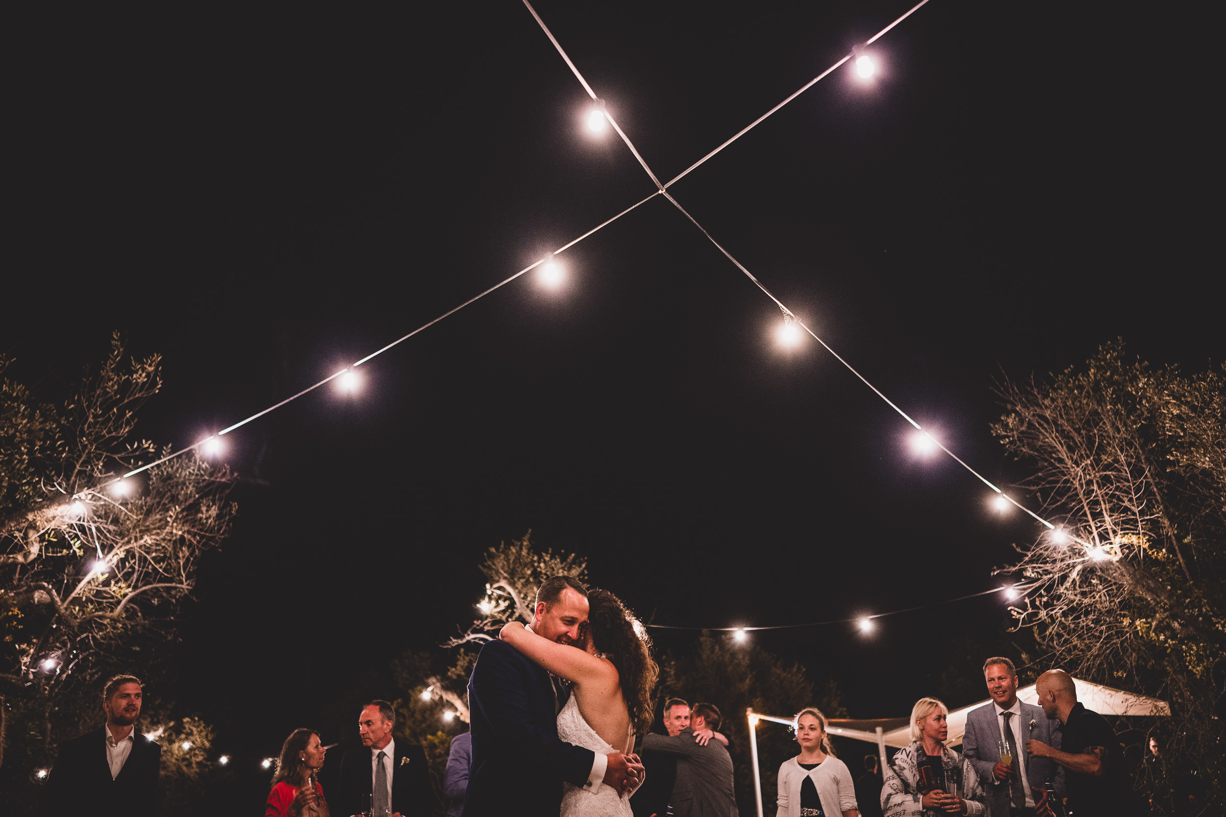 A wedding photo of the bride and groom kissing under string lights at night.