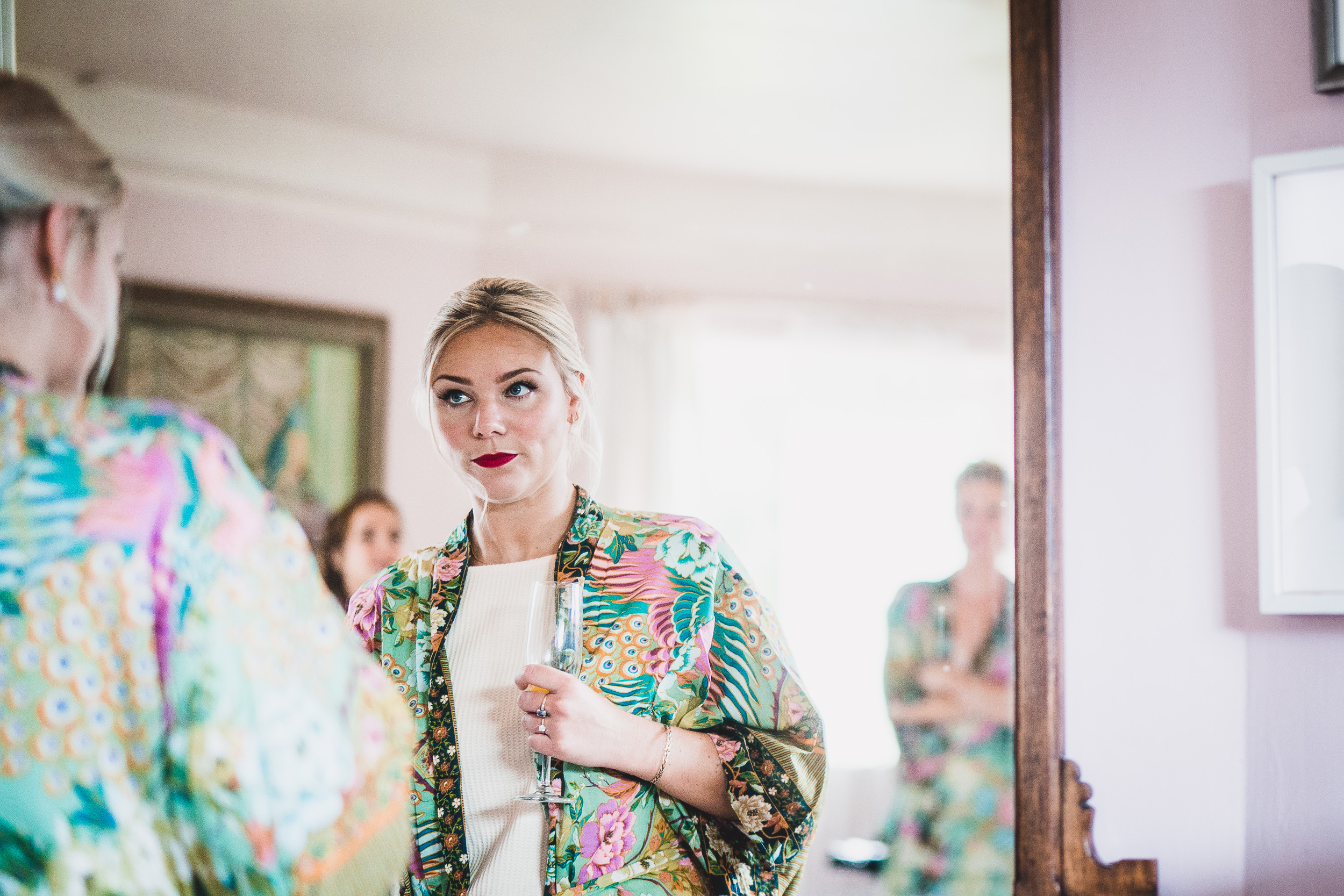 A bride in a floral robe is admiring herself in the mirror, with the help of a wedding photographer capturing the moment.