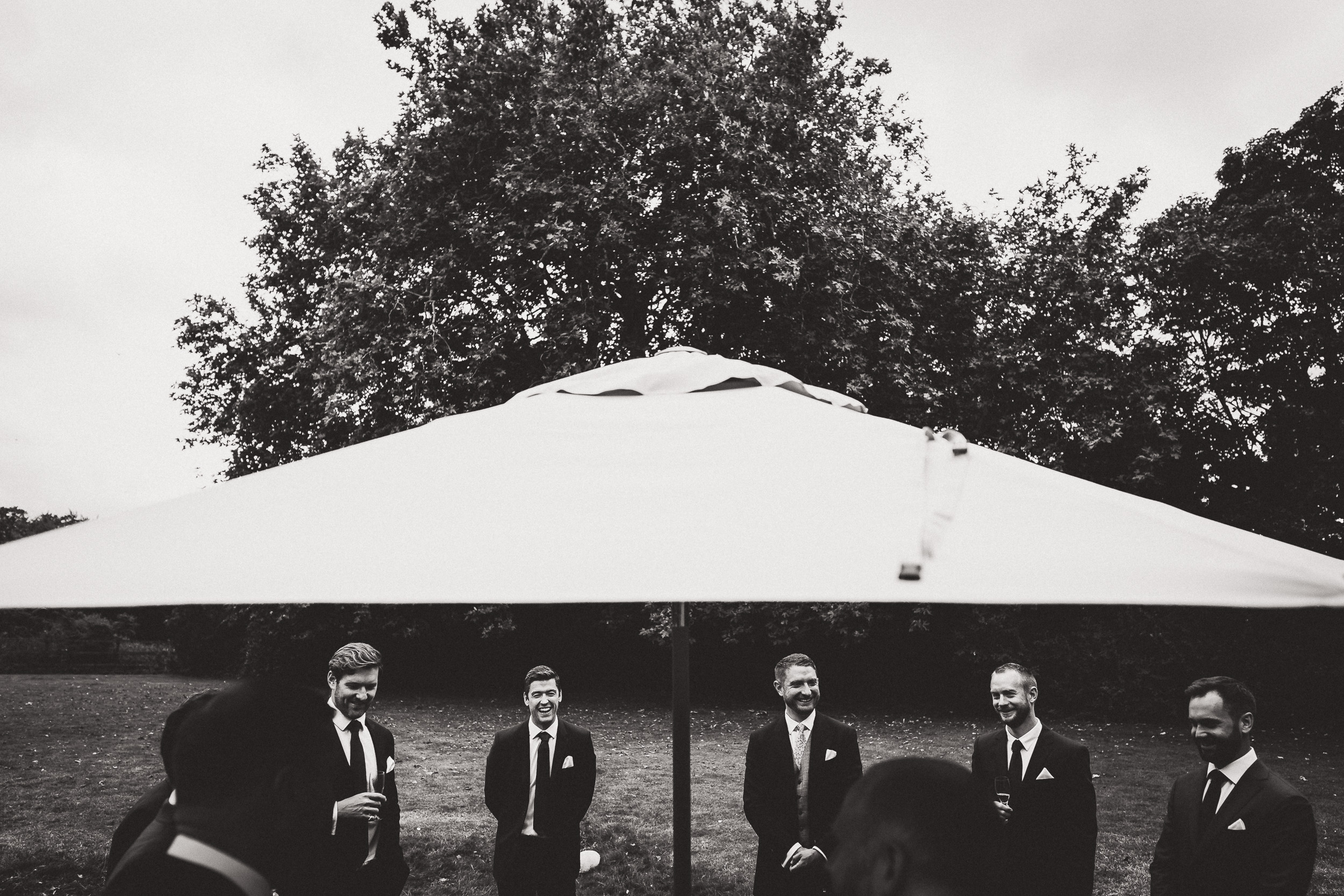 A group of groomsmen standing under an umbrella at a wedding.