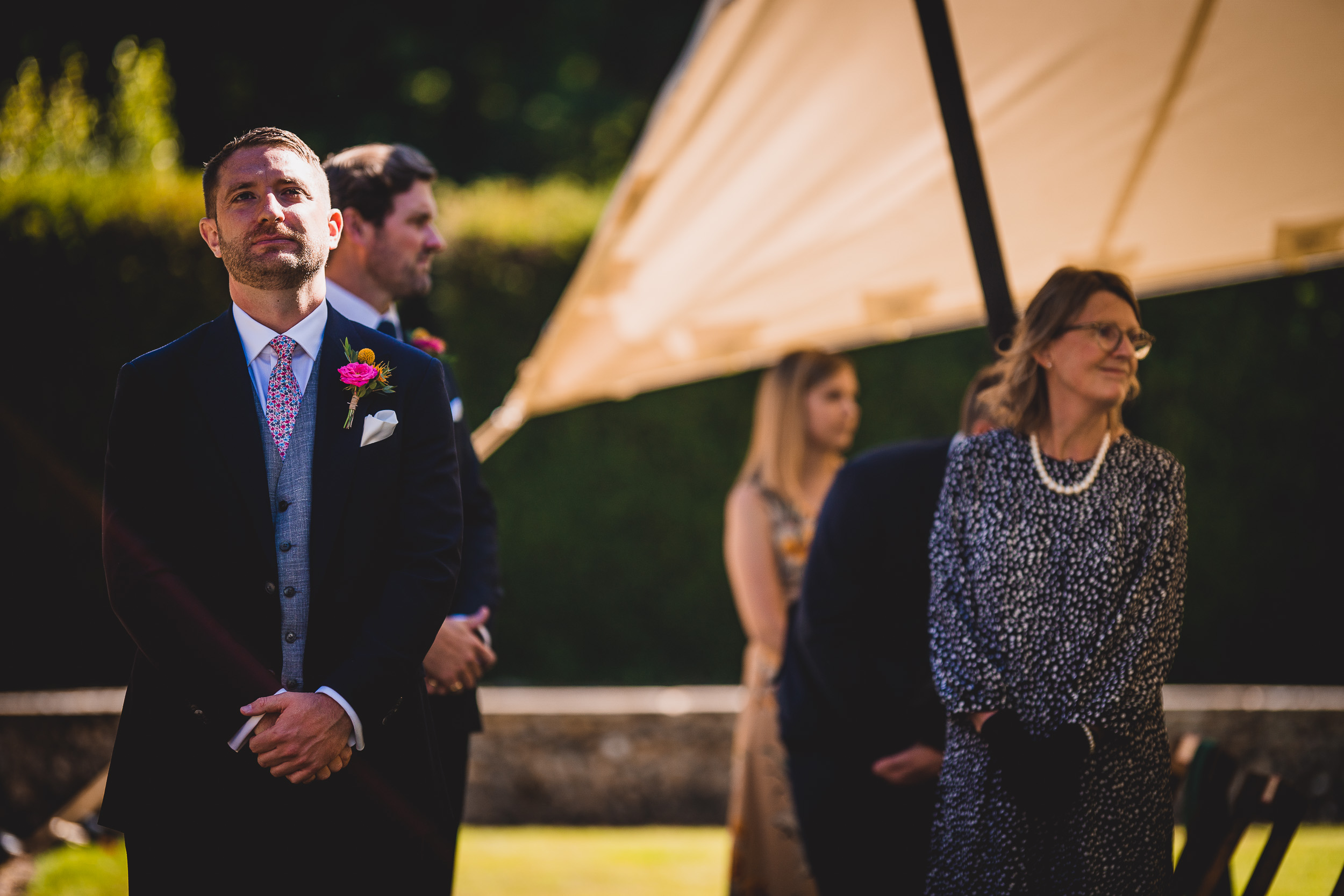 A groom in a suit is standing in front of a wedding tent.