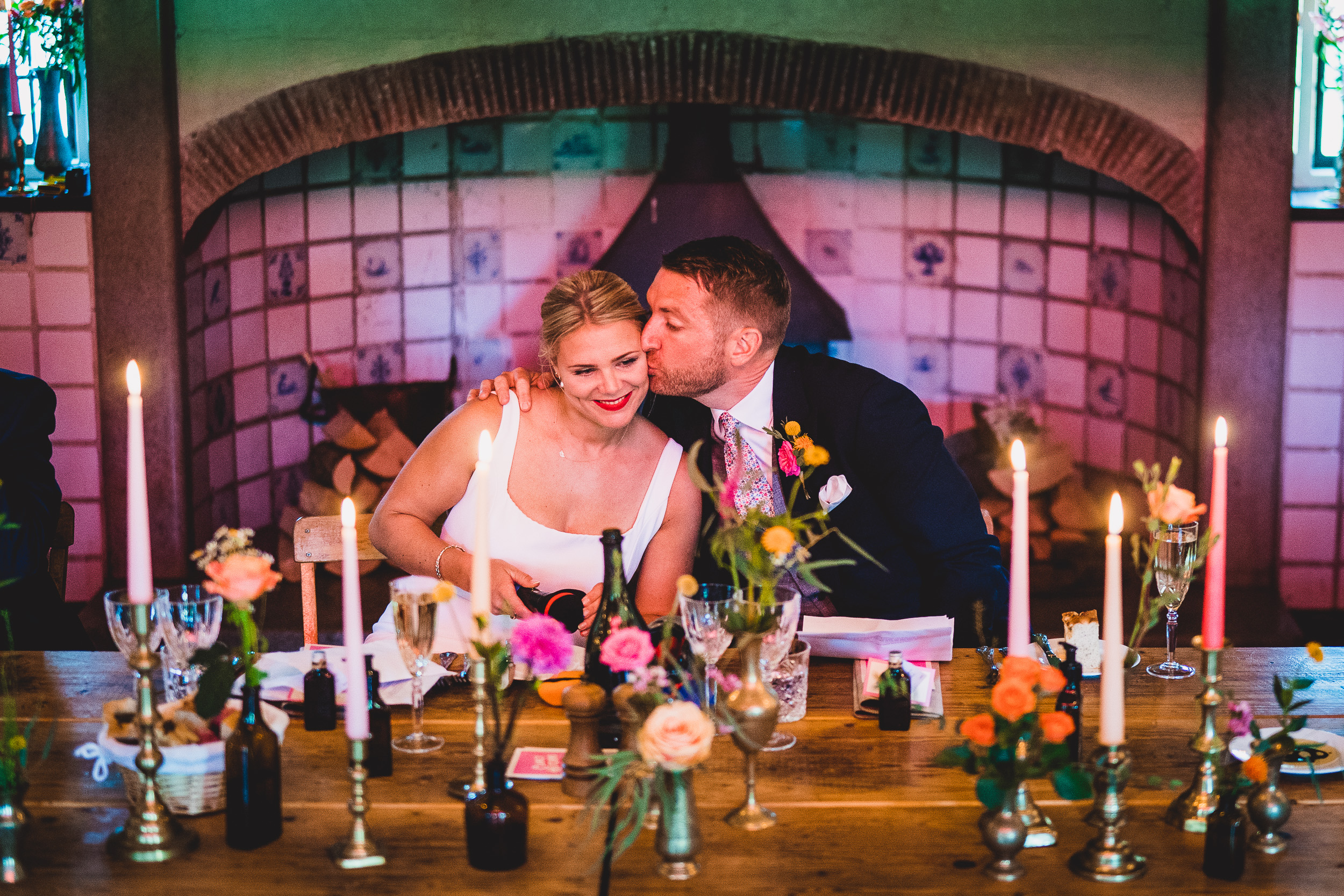 A wedding photographer captures a romantic moment of the groom kissing his bride at a table illuminated by candles.