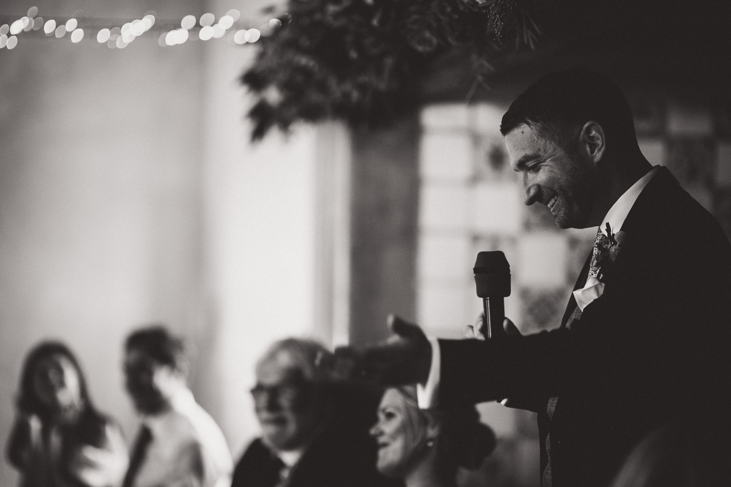 A black and white wedding photo capturing the groom giving a speech.