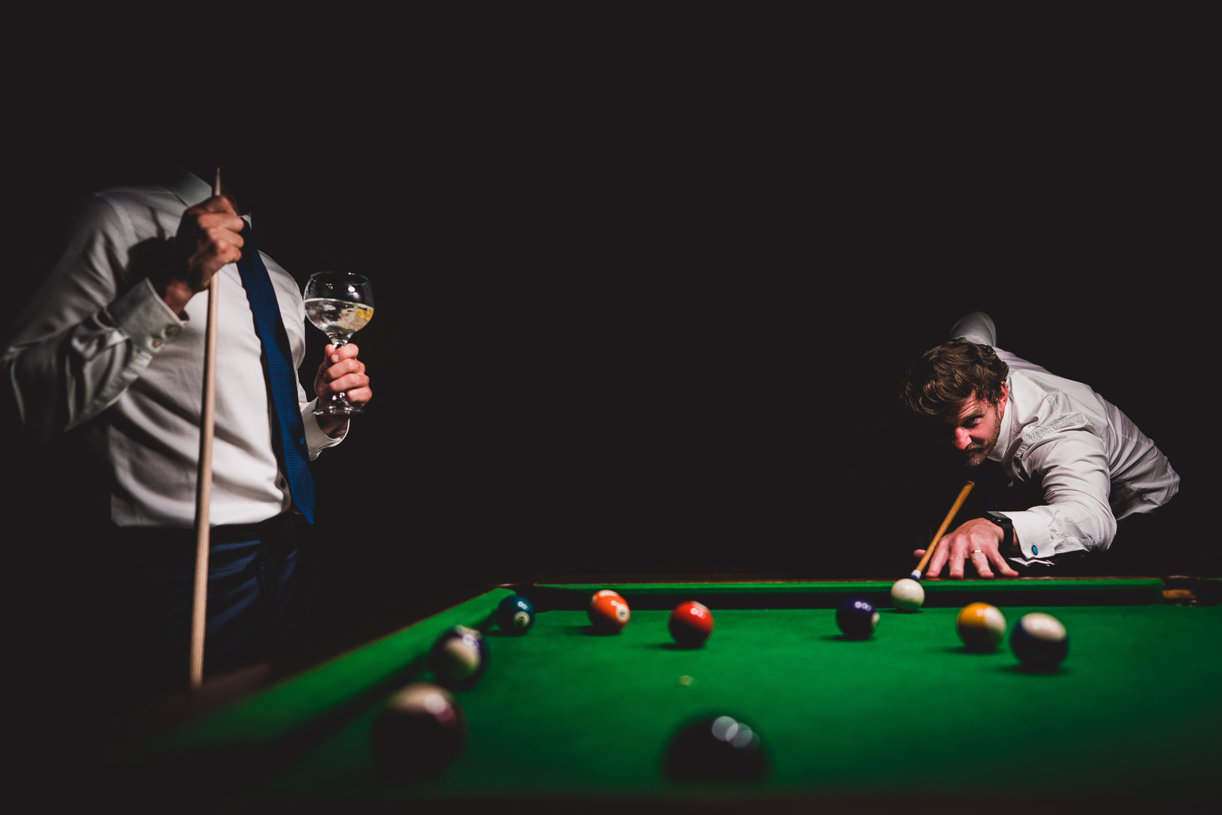 A groom is playing snooker with a glass of wine.