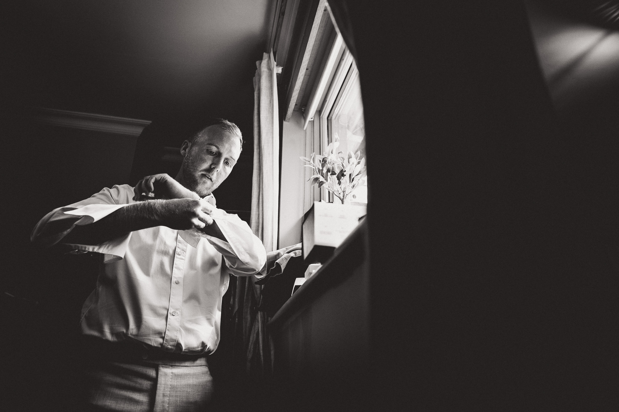 A groom is adjusting his watch in front of a window on his wedding day.