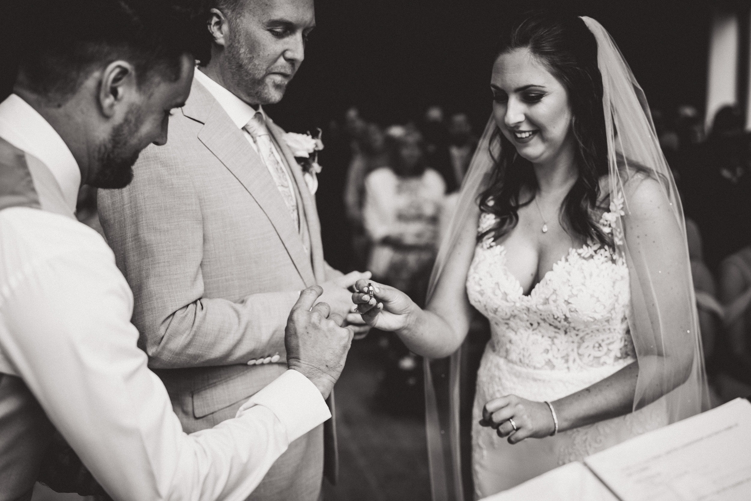 A bride and groom exchange rings during their wedding ceremony while being captured by a wedding photographer.