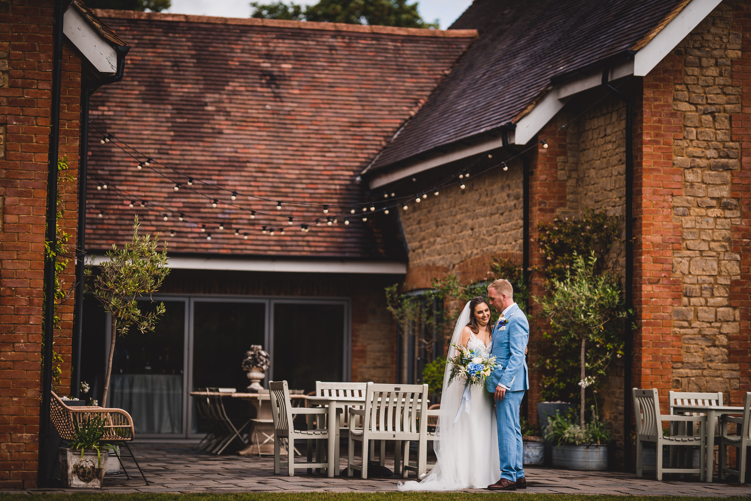 A groom and bride posing for the wedding photographer in front of a brick building.