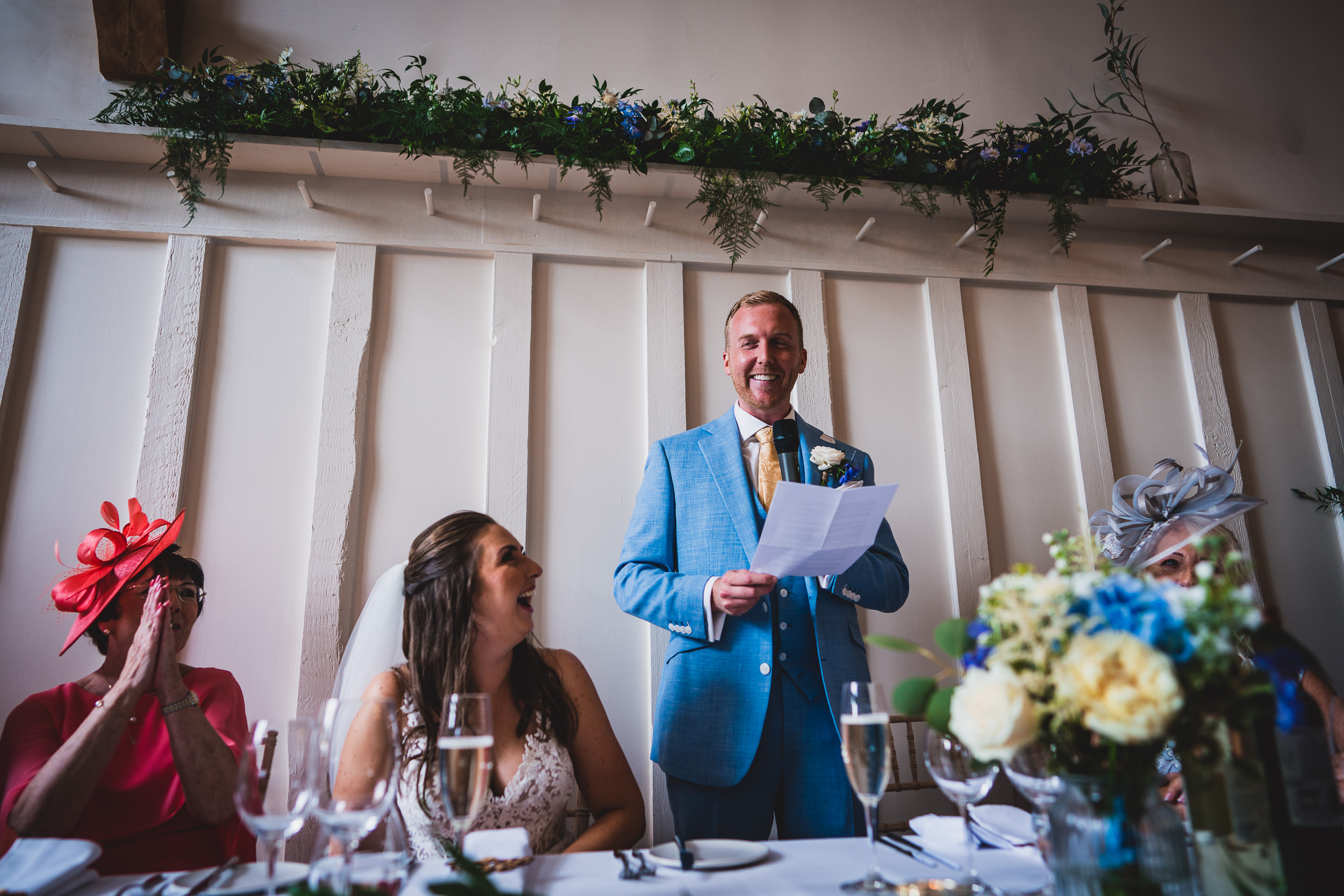 A wedding photographer captures the moment of a man in a blue suit giving a speech at the bride's wedding.