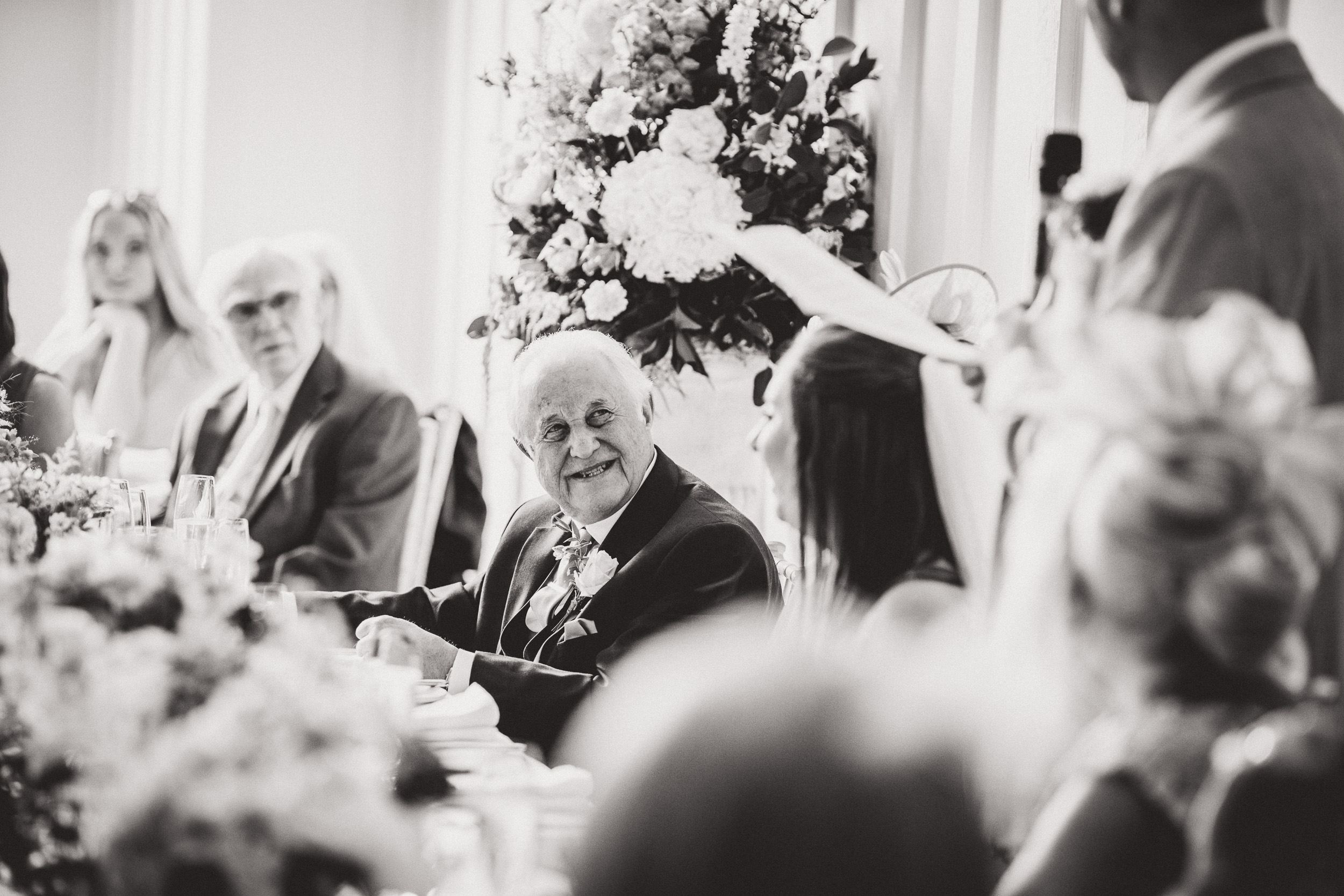 A black and white wedding photo of a man speaking at the reception.