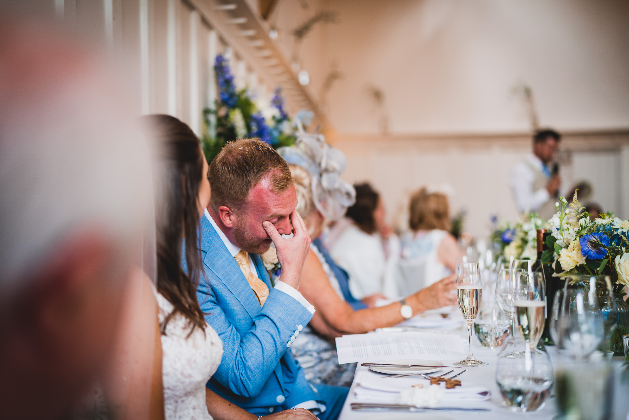 The wedding photographer captures a tearful man in a blue suit at the reception.