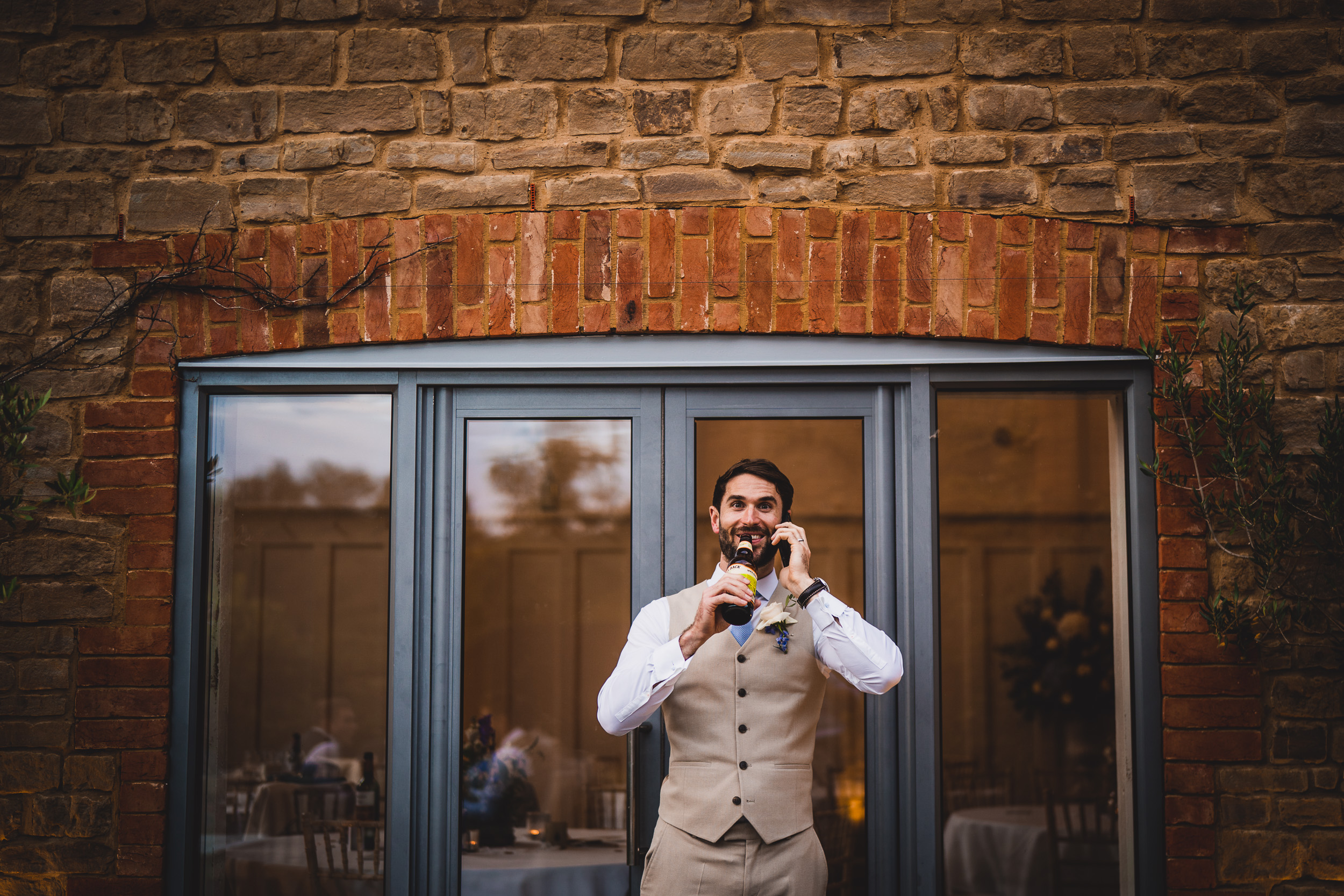 A groom talking on a cell phone in front of a door with a wedding photographer.