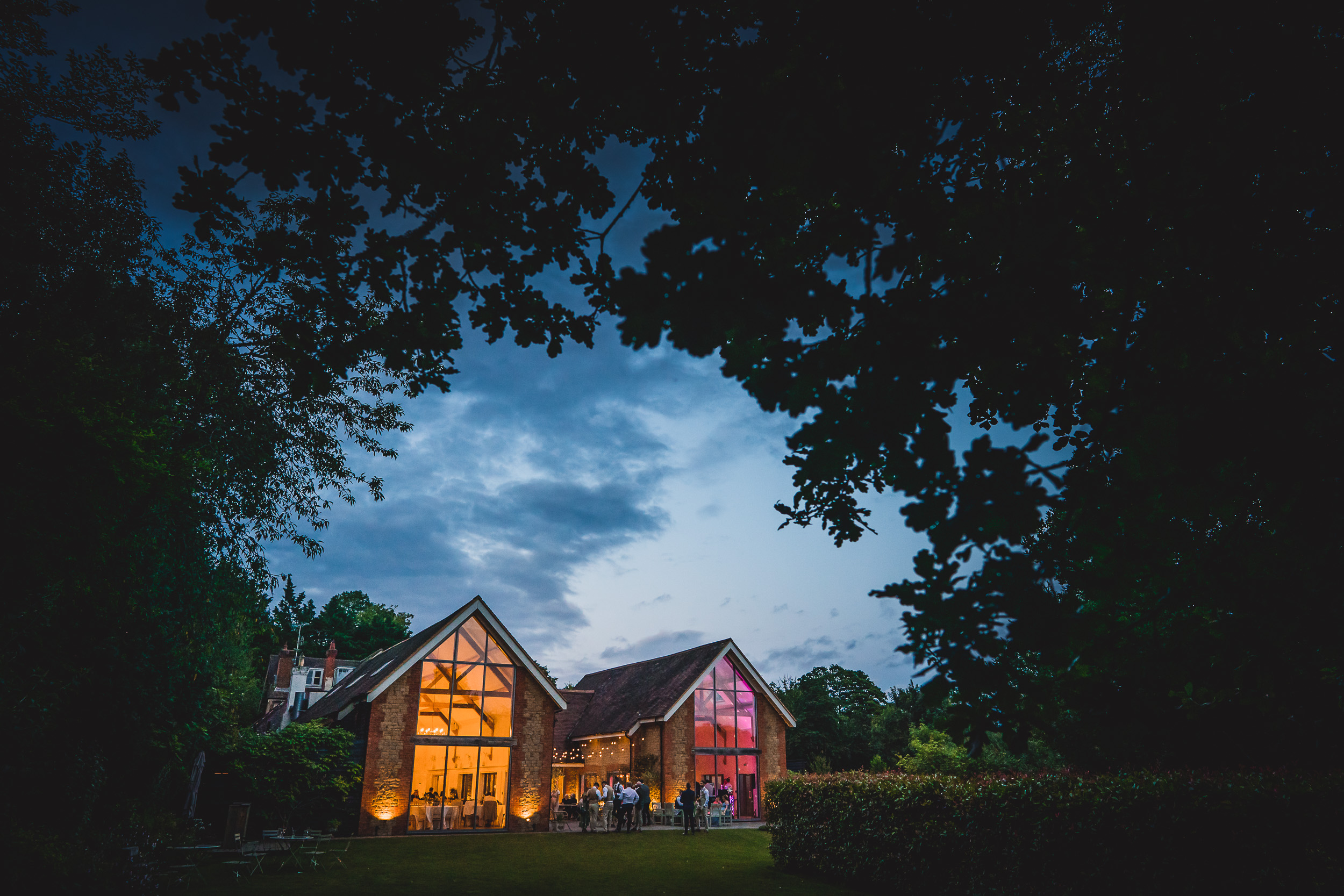 A wedding photo of a groom outside a house lit up at dusk in a wooded area.