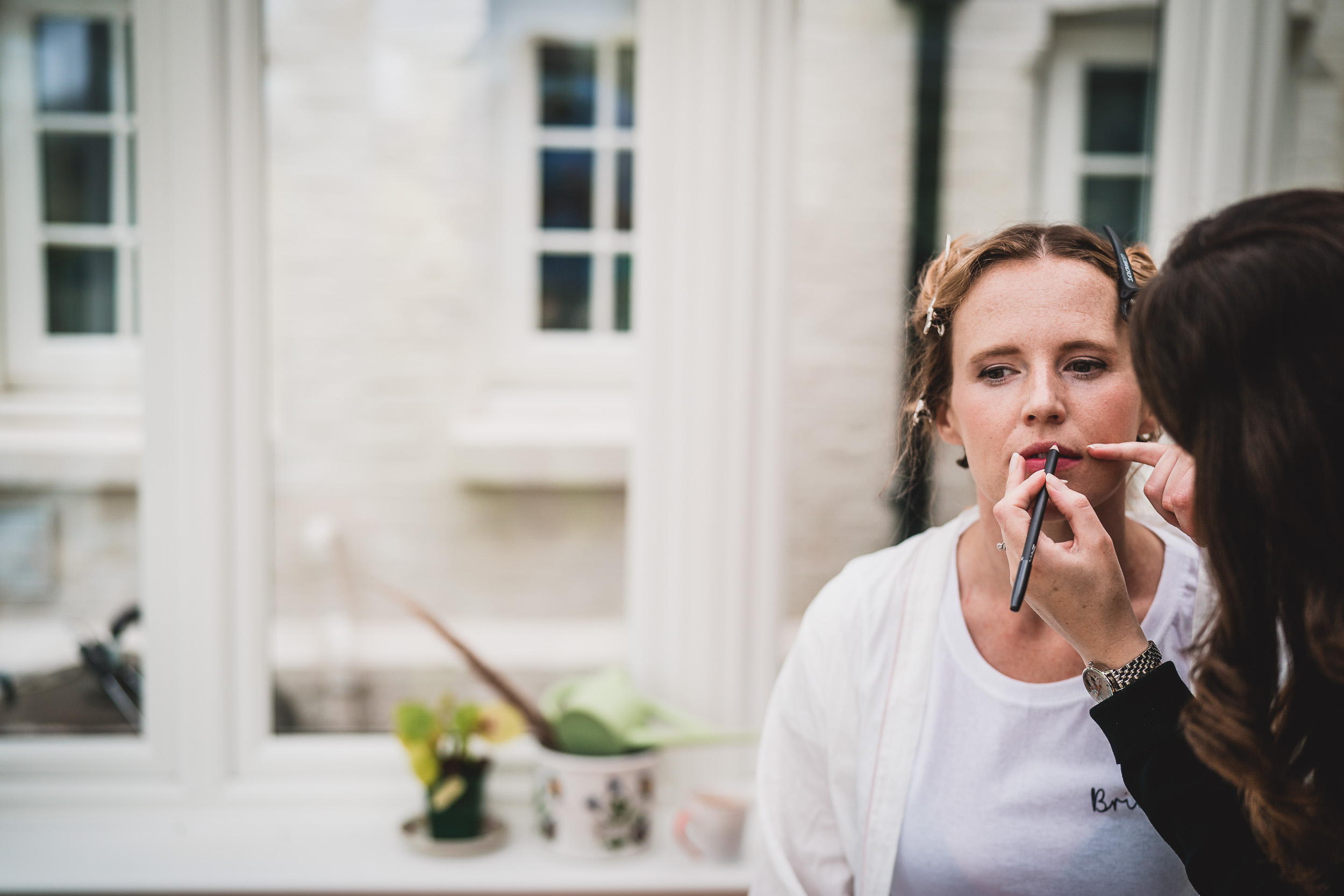 A bride getting her makeup done in front of a window.