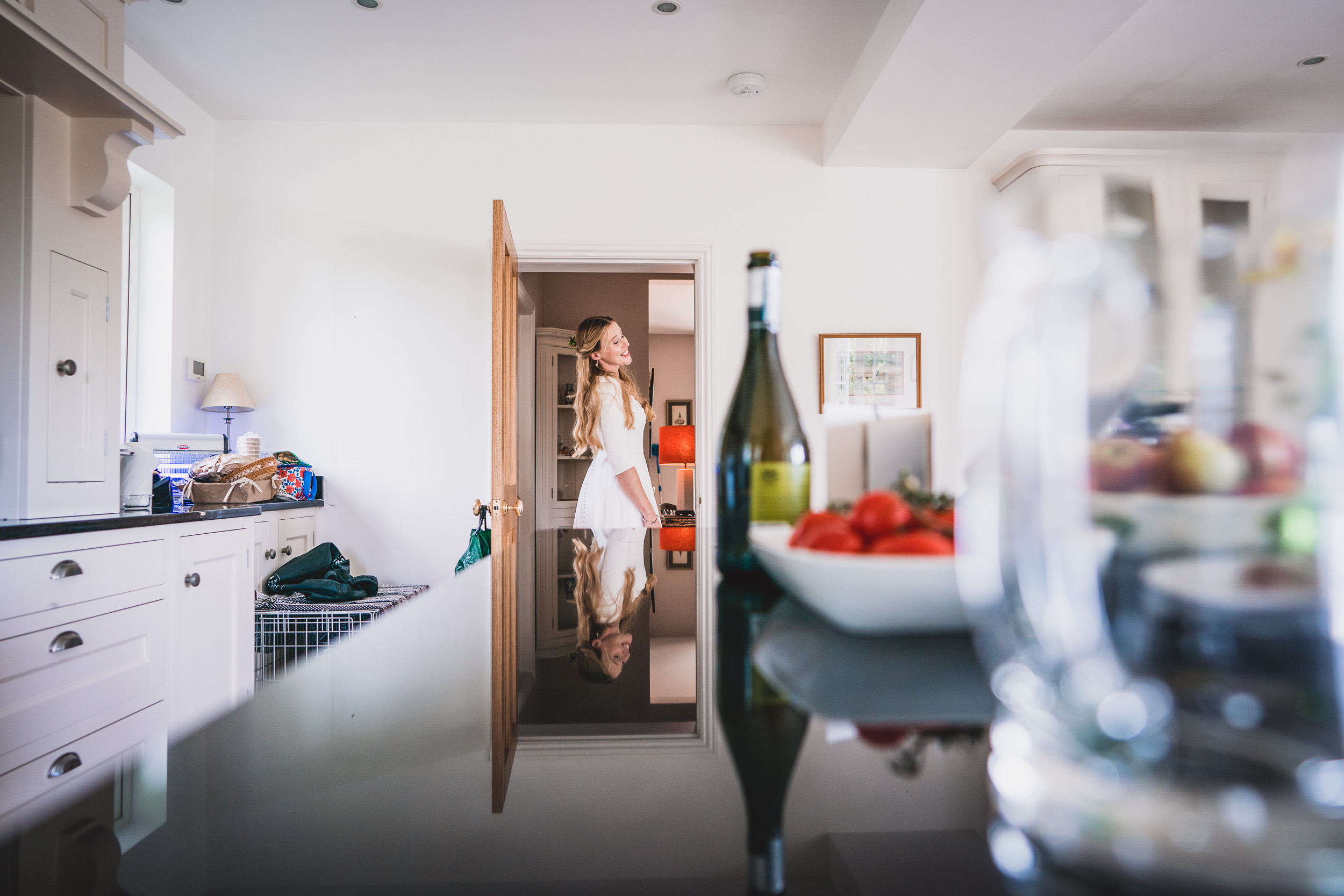 A bride in a white dress standing in a kitchen being photographed by a wedding photographer.