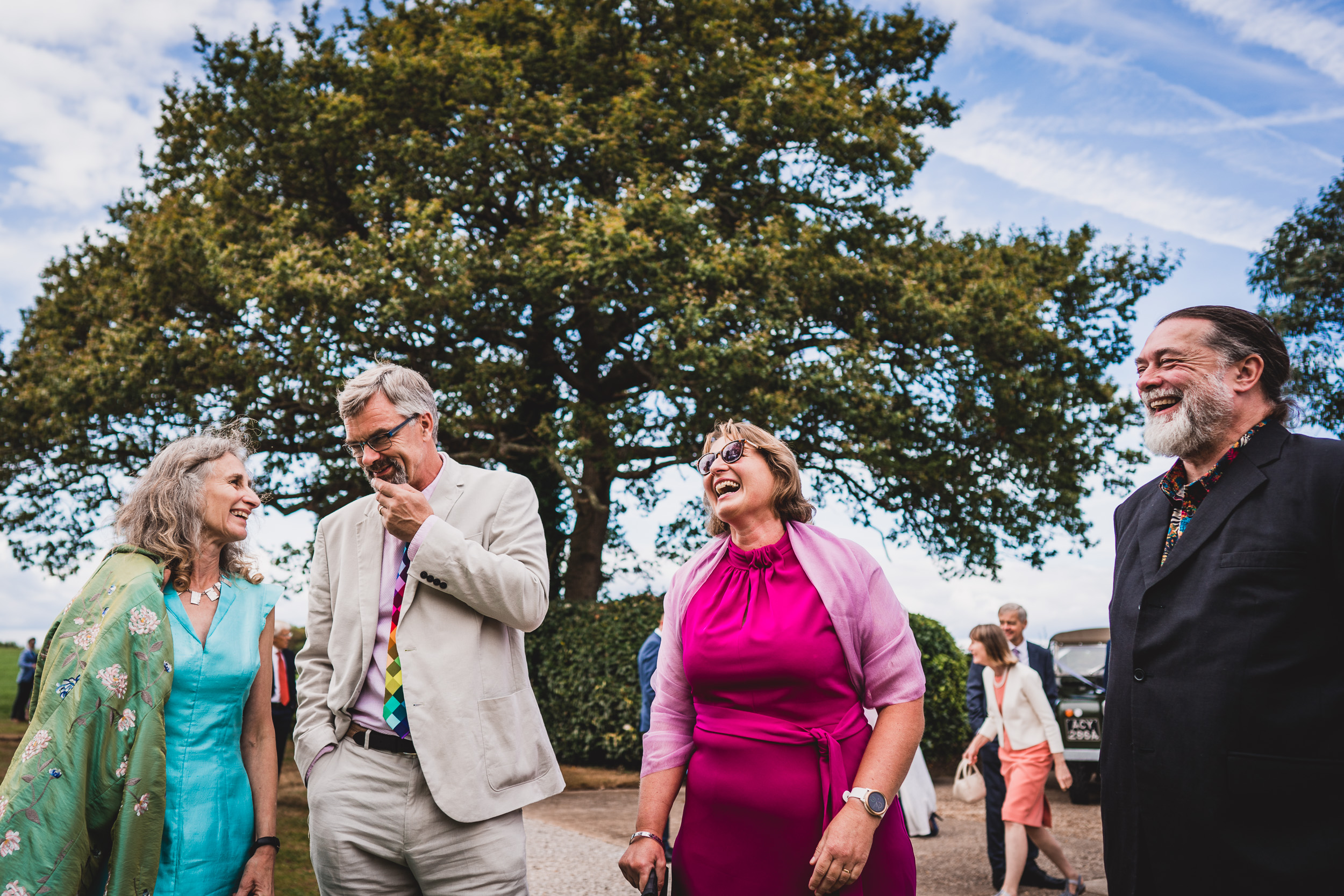 A group of people, including the bride and groom, laughing in front of a tree captured by the wedding photographer.