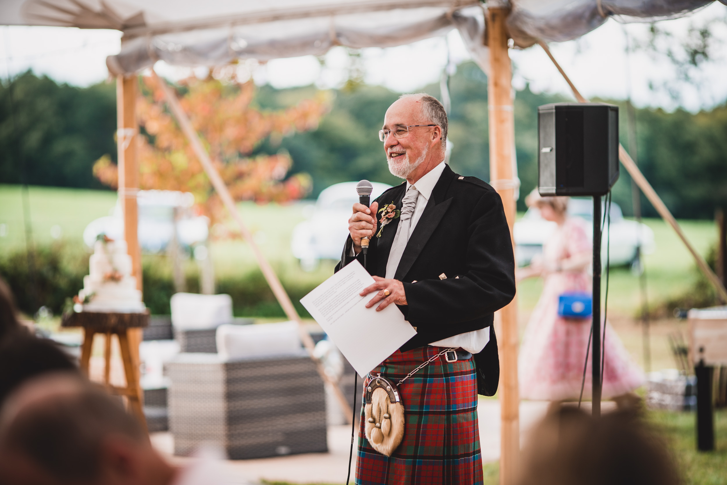 A groom in a kilt speaking into a microphone at his wedding.