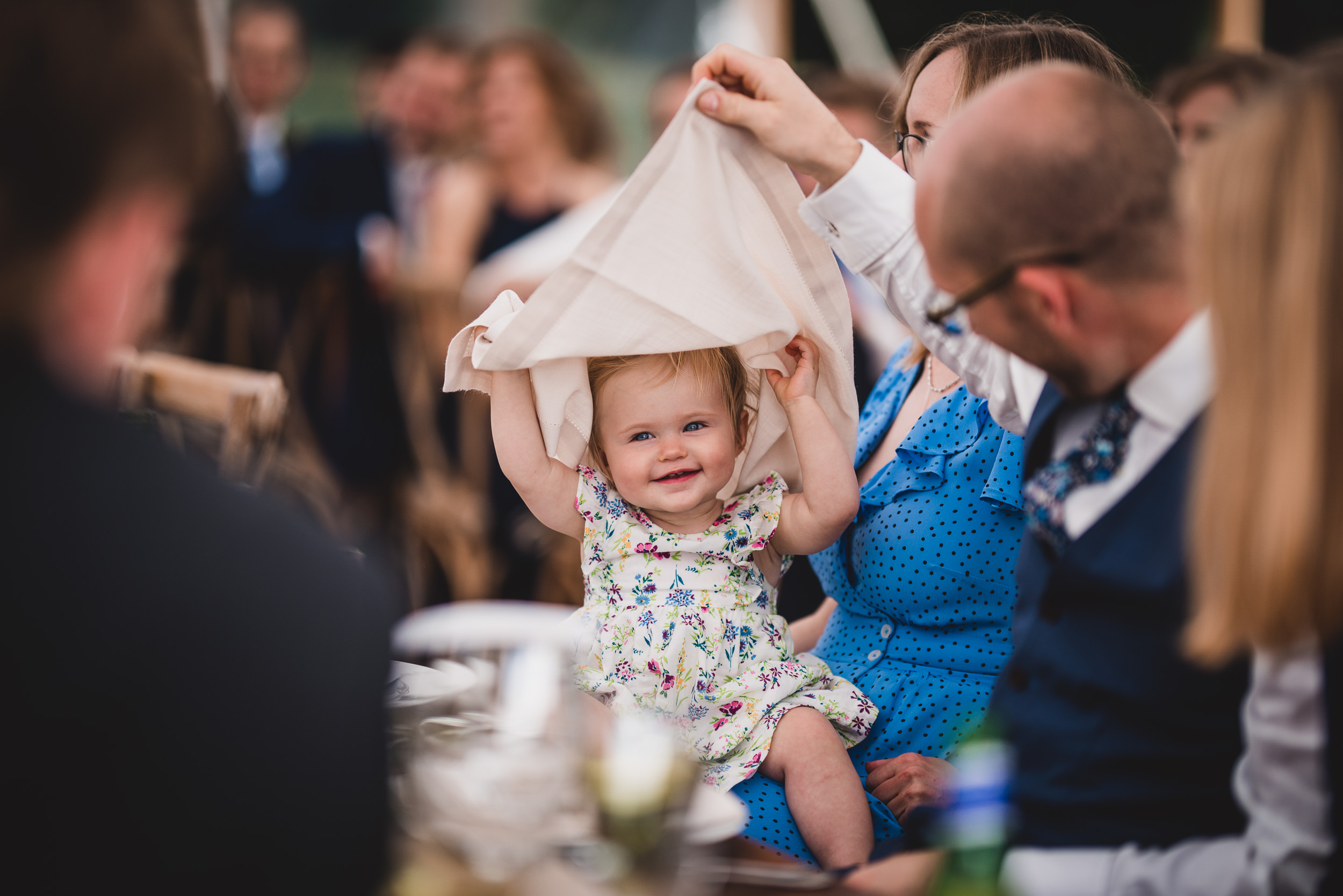 A little girl is holding up a napkin at a wedding reception, disrupting the wedding photo.