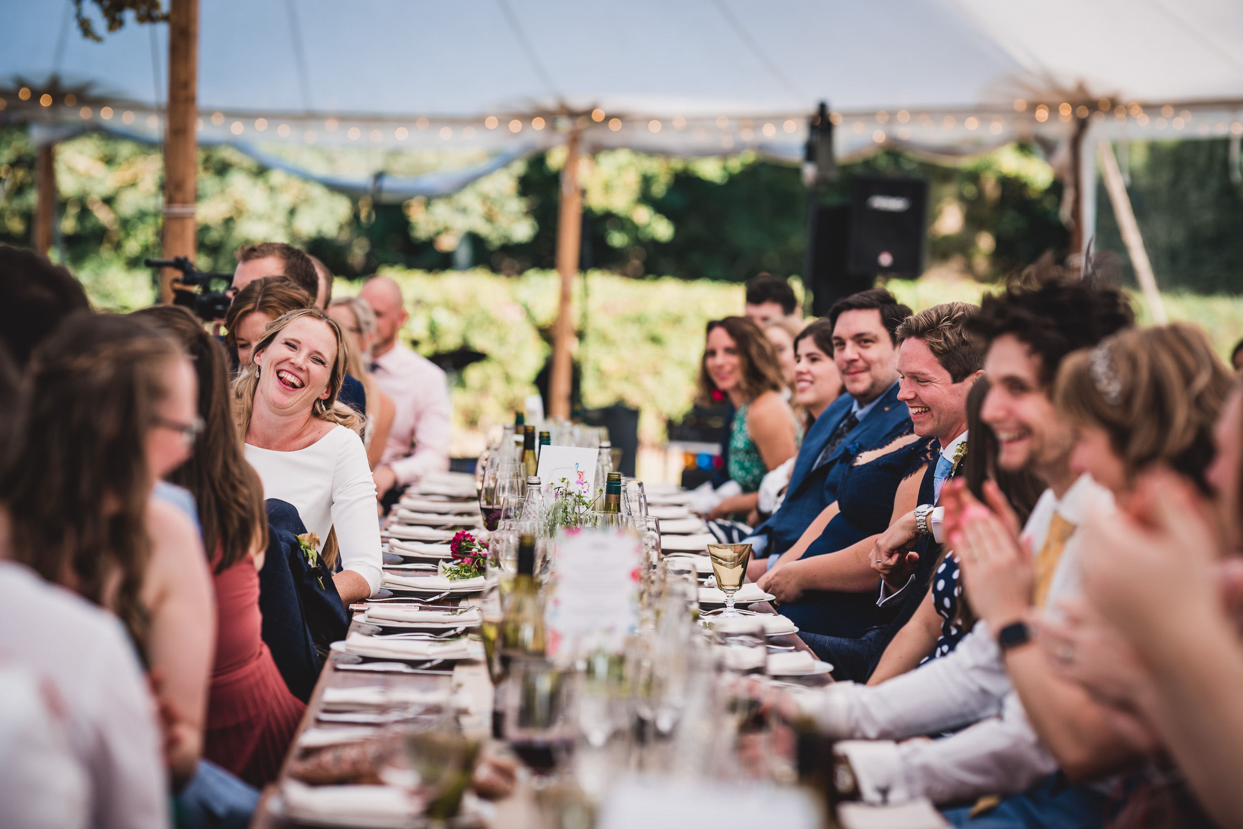 A wedding photo capturing the groom and bride along with their guests sitting at a long table in a tent.
