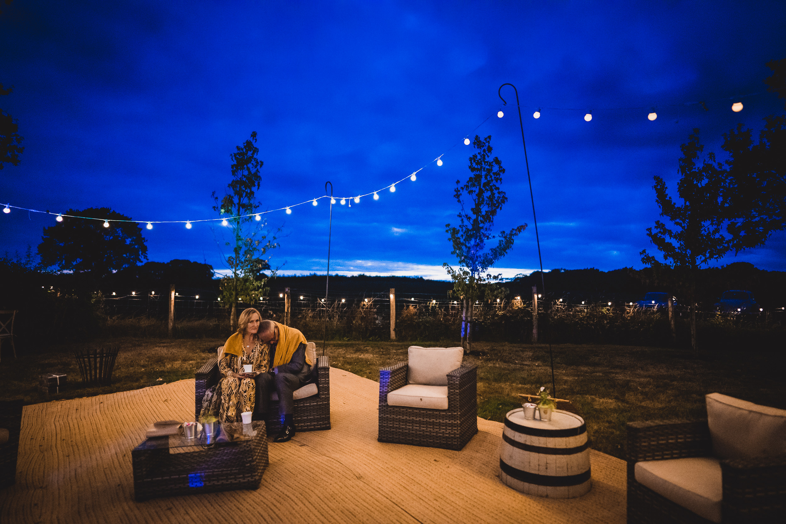 A bride and groom sit on a patio under string lights at dusk, captured by their wedding photographer.