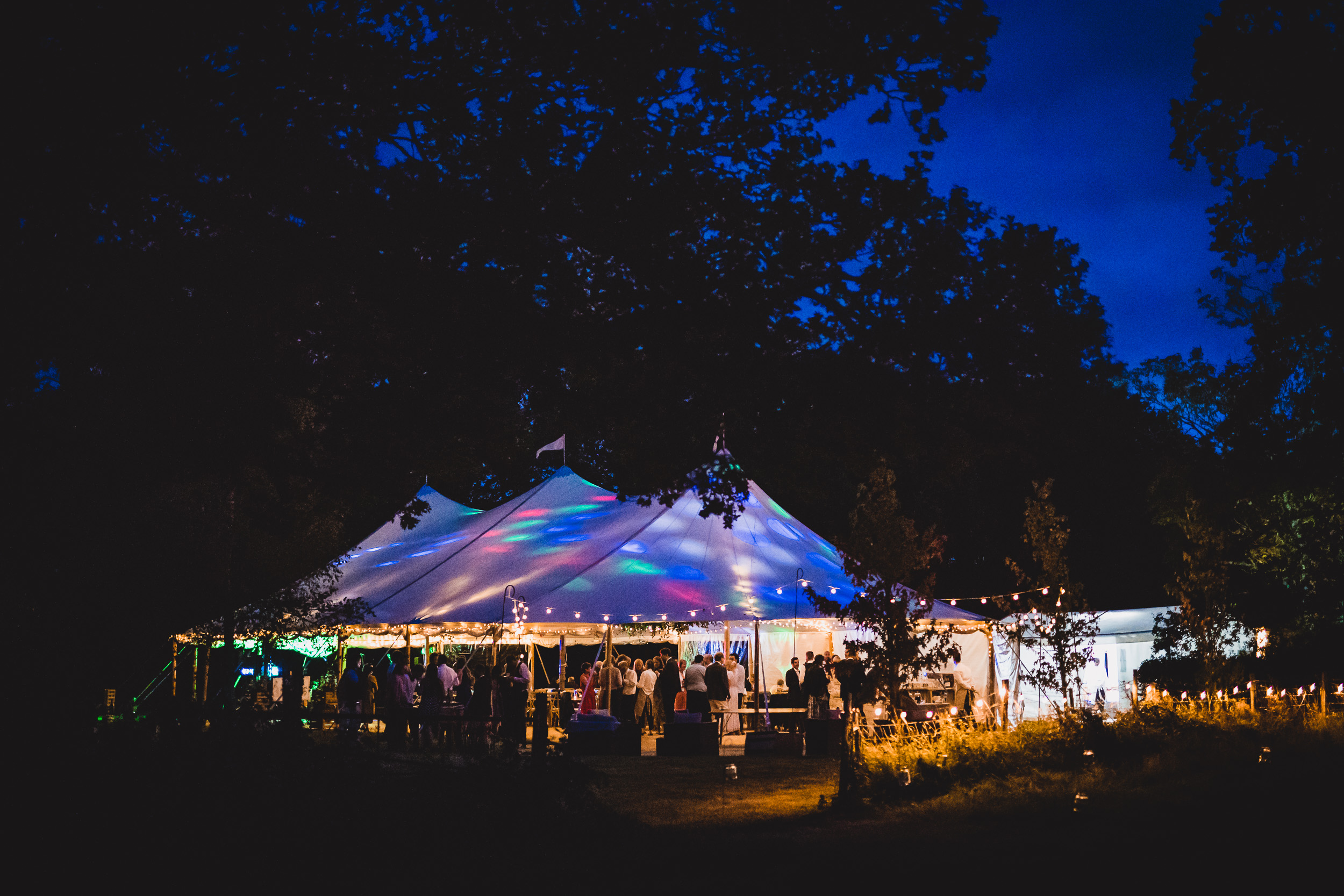 A bride stands outside a tent lit up at night in a wooded area, posing for her wedding photo.