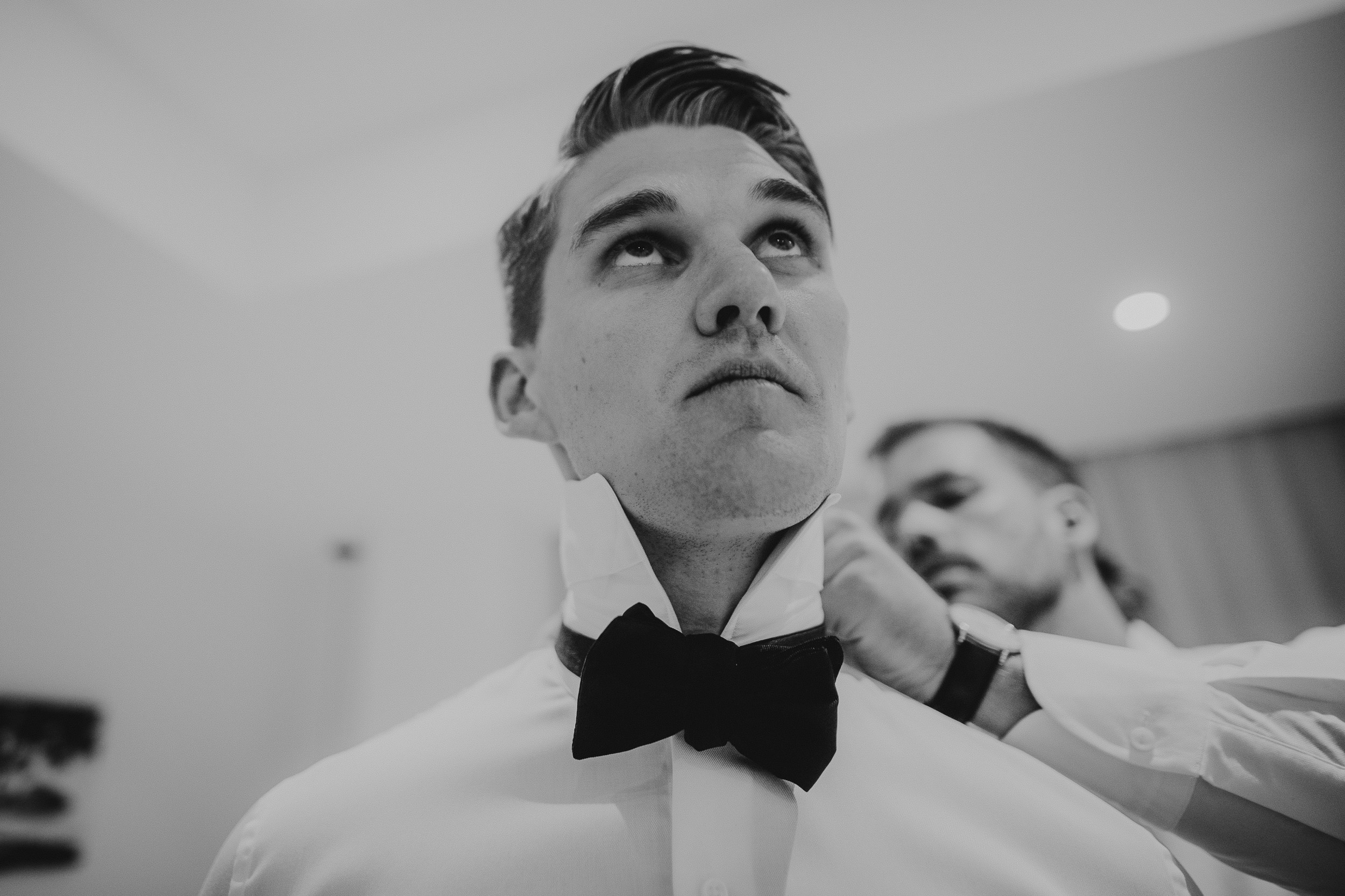 A groom is adjusting his bow tie in a black and white wedding photo.