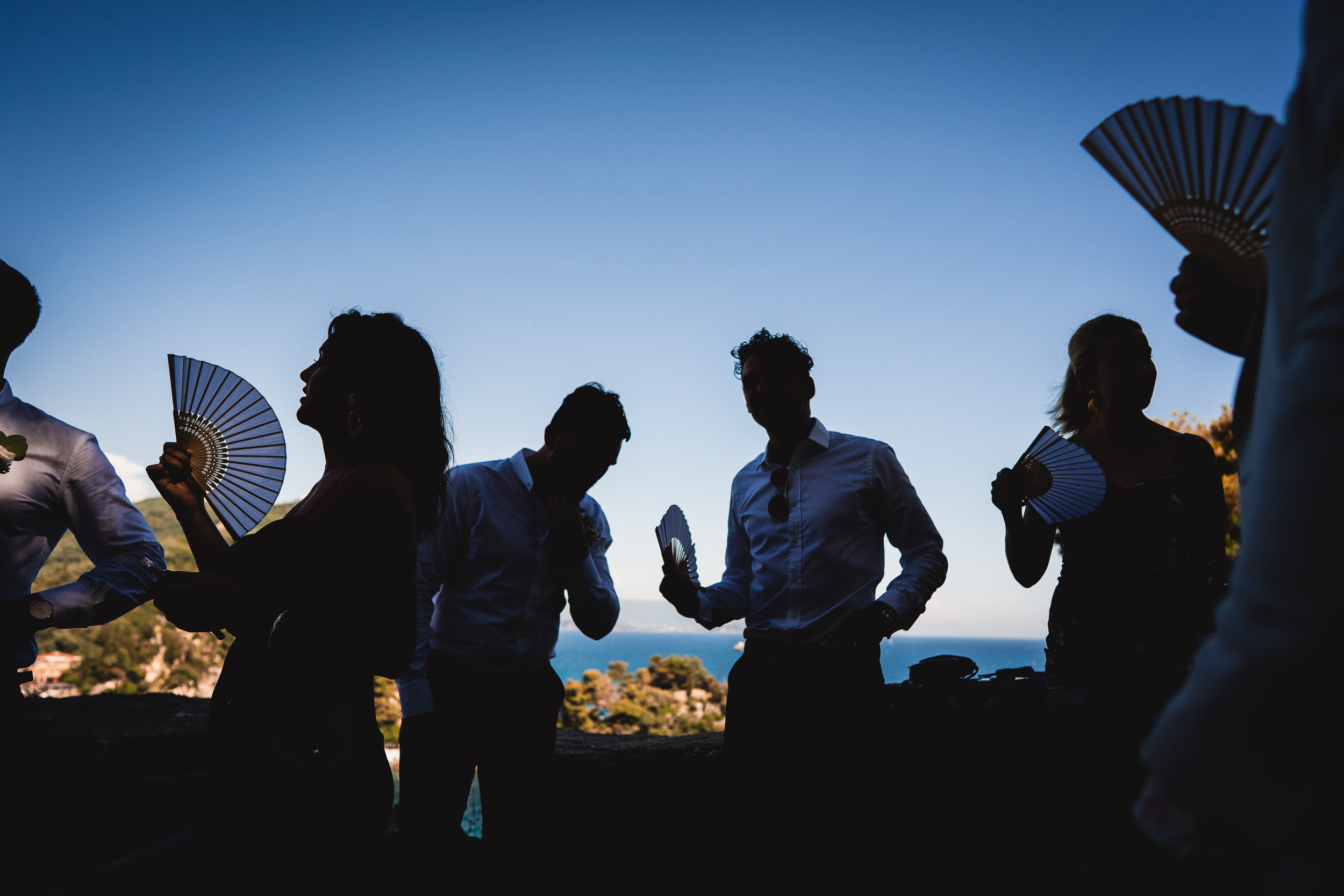 A wedding photographer capturing a bride and group of people holding fans by the ocean.