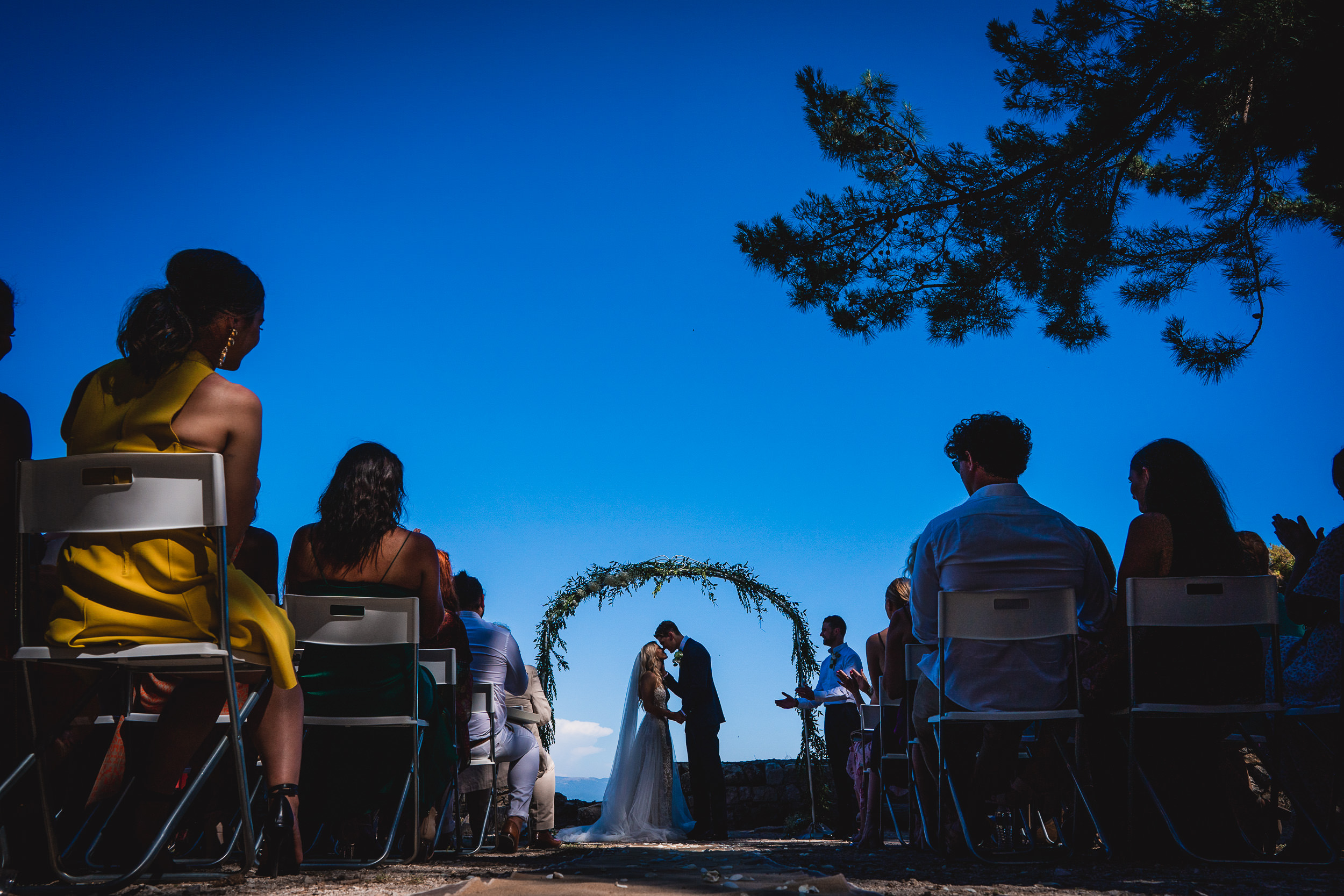 A groom and bride posing for their wedding photographer under a blue sky.
