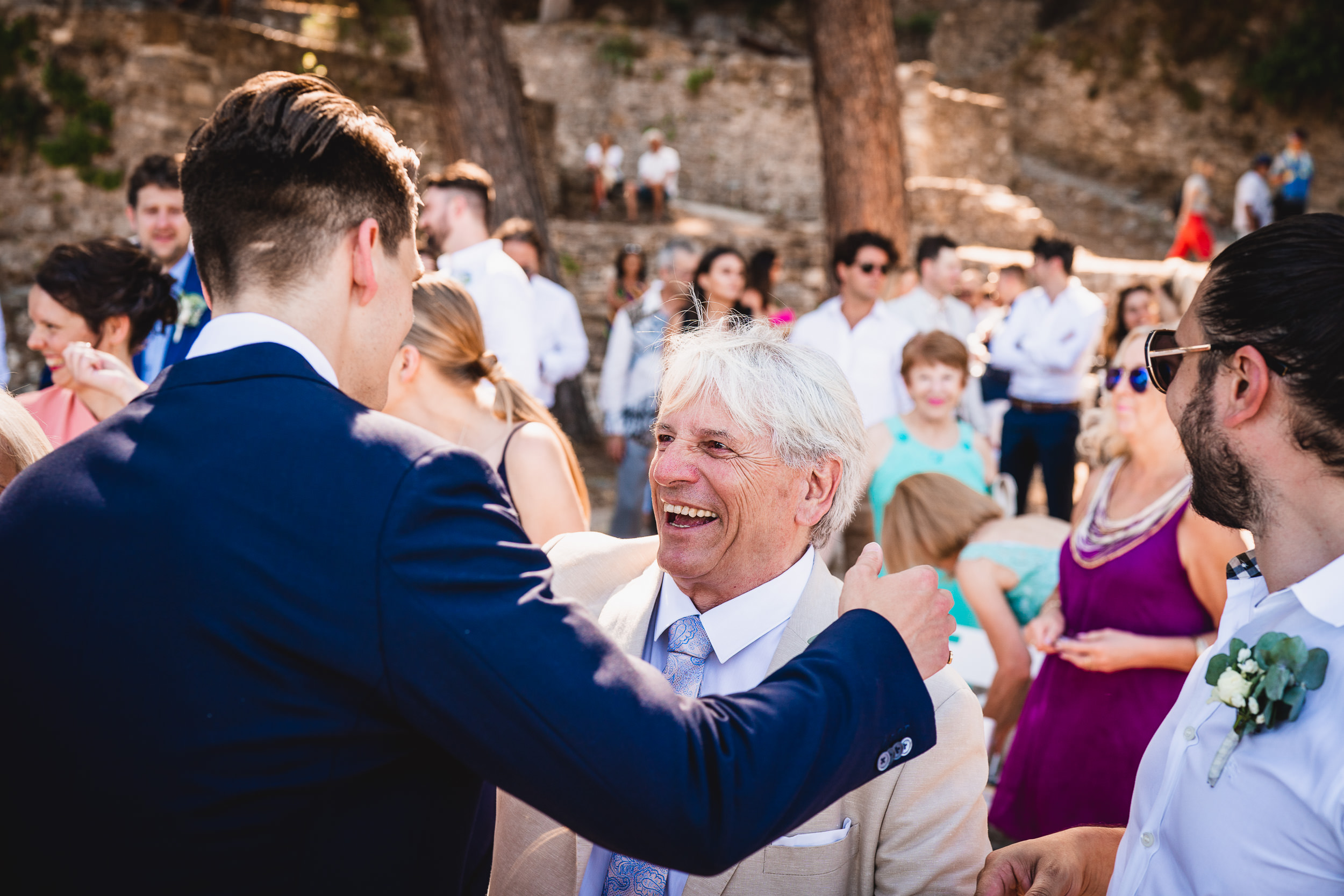 At a wedding, a man in a suit is laughing with another man while the wedding photographer captures the moment.