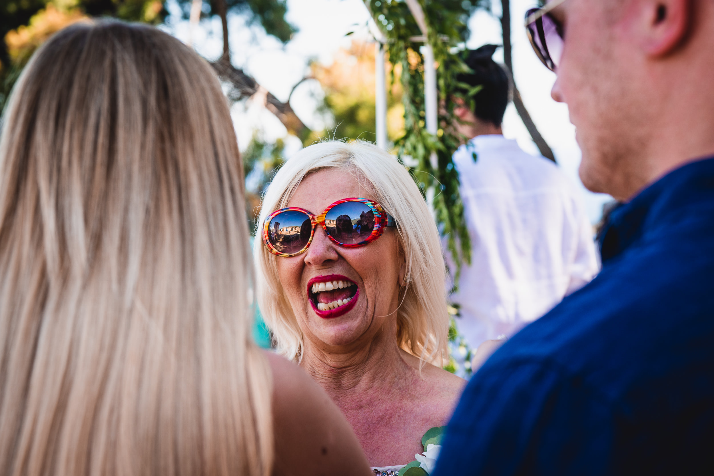 A bride wearing sunglasses is talking to the groom at an outdoor wedding photo shoot.