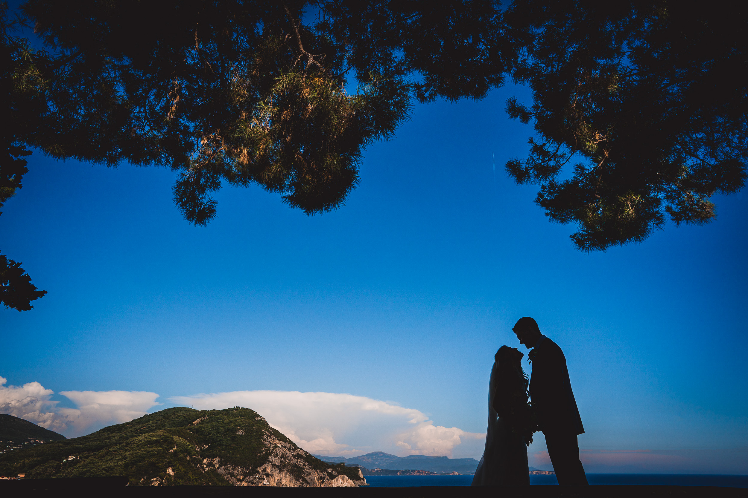 A groom and bride pose for their wedding photographer in front of the ocean.