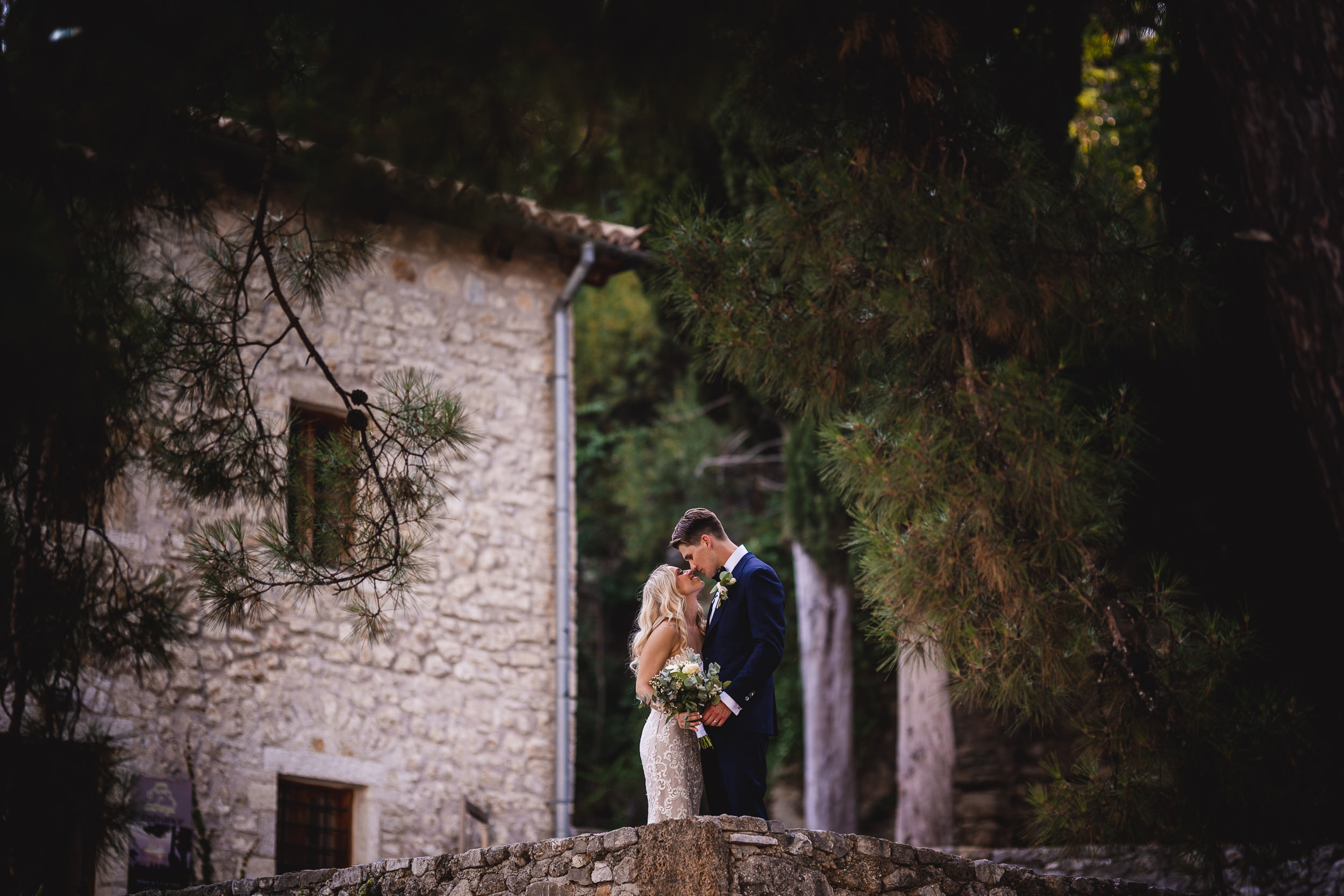 A wedding couple posing on a stone wall near a stone house.