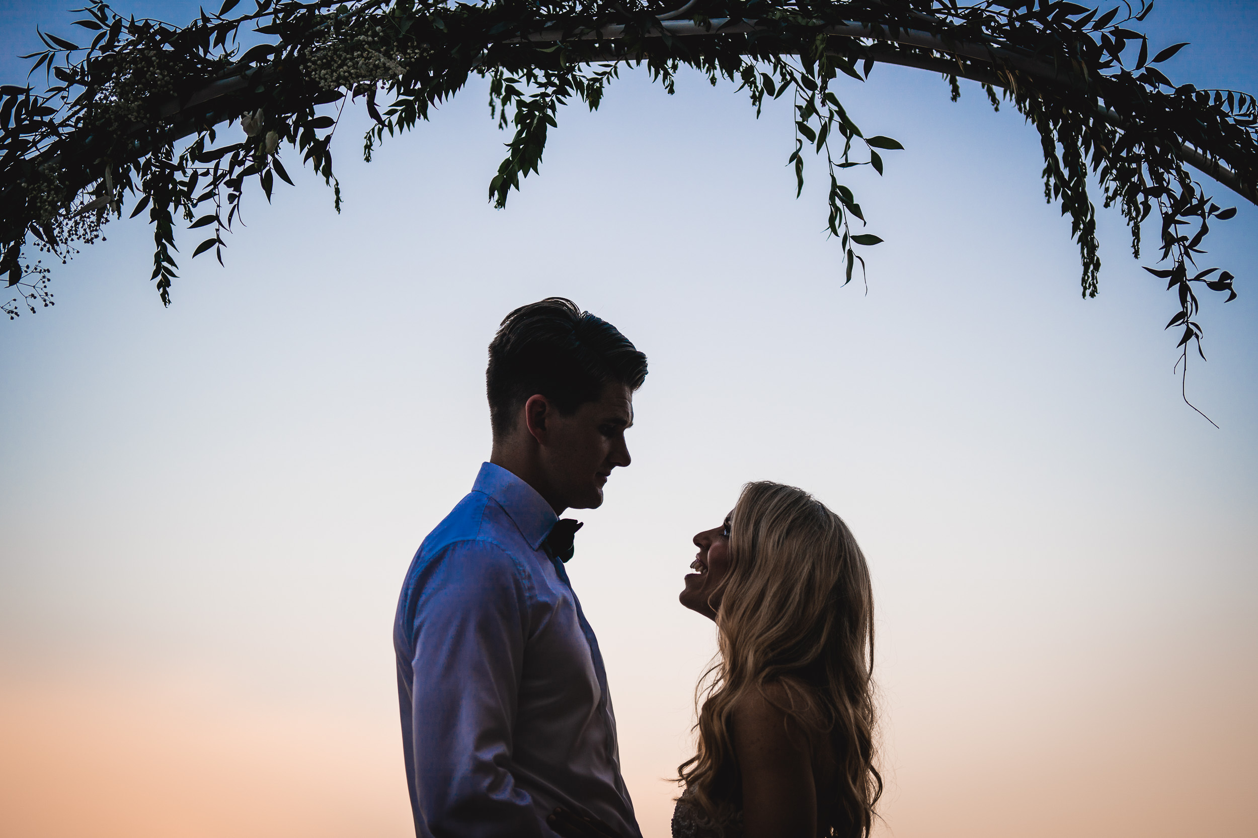 A wedding photo capturing the silhouette of a bride and groom standing under an arch at sunset.