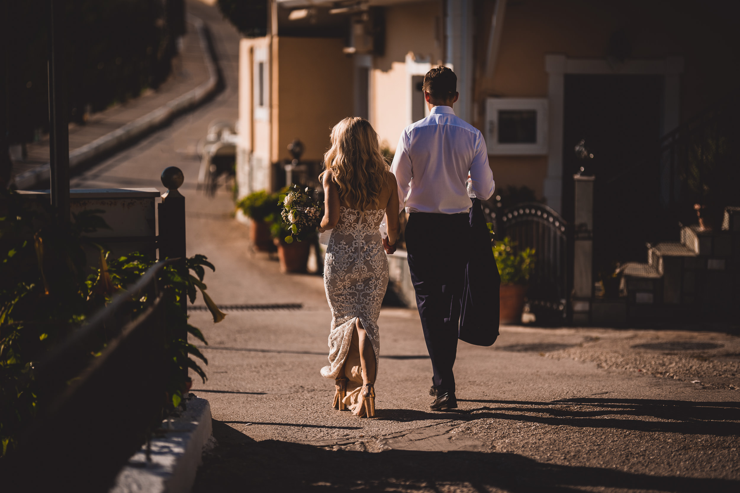 A wedding photographer captures the groom and bride strolling down a narrow street on their special day.