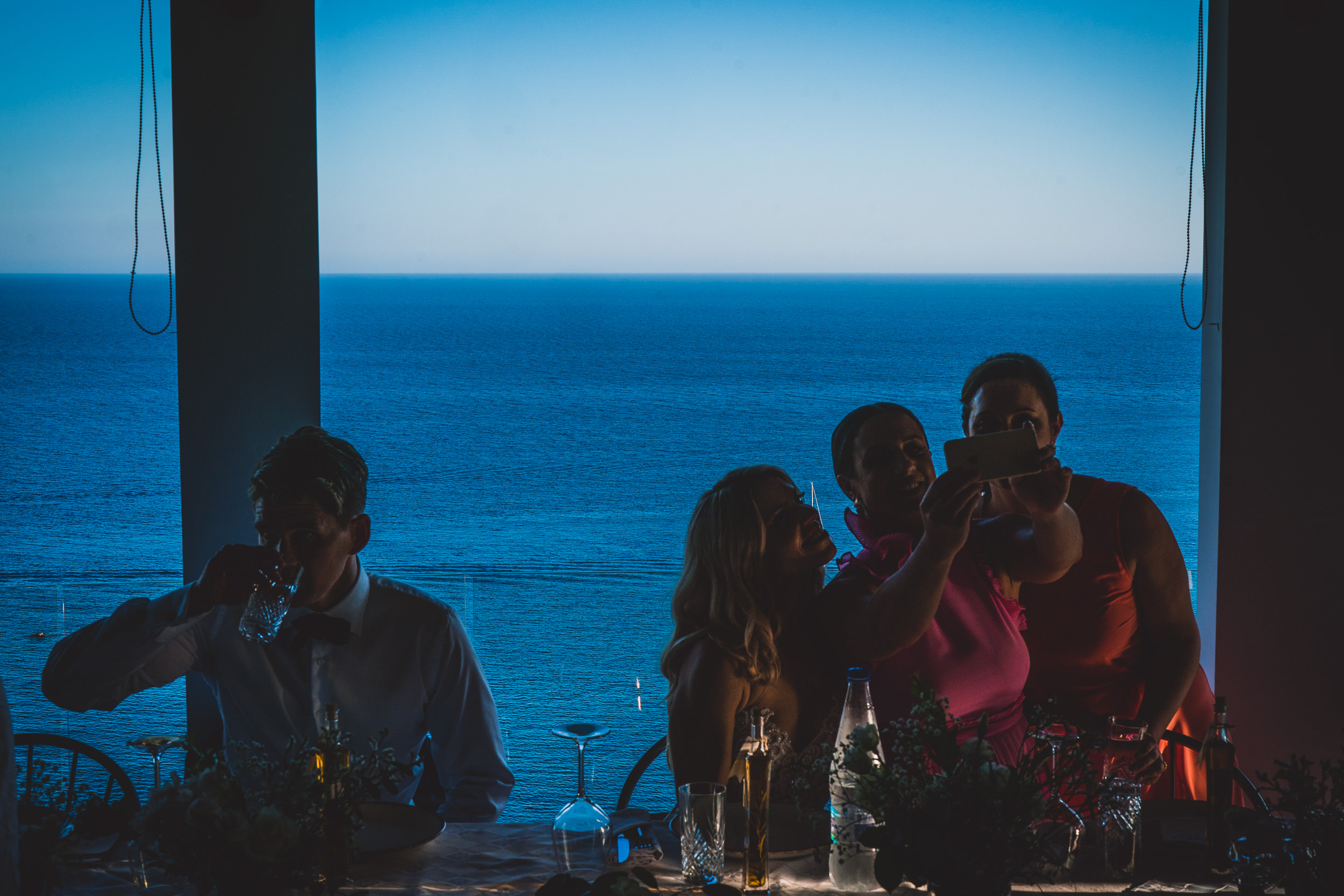 A bride and her wedding photographer capturing a picture-perfect moment at a table with a view of the ocean.