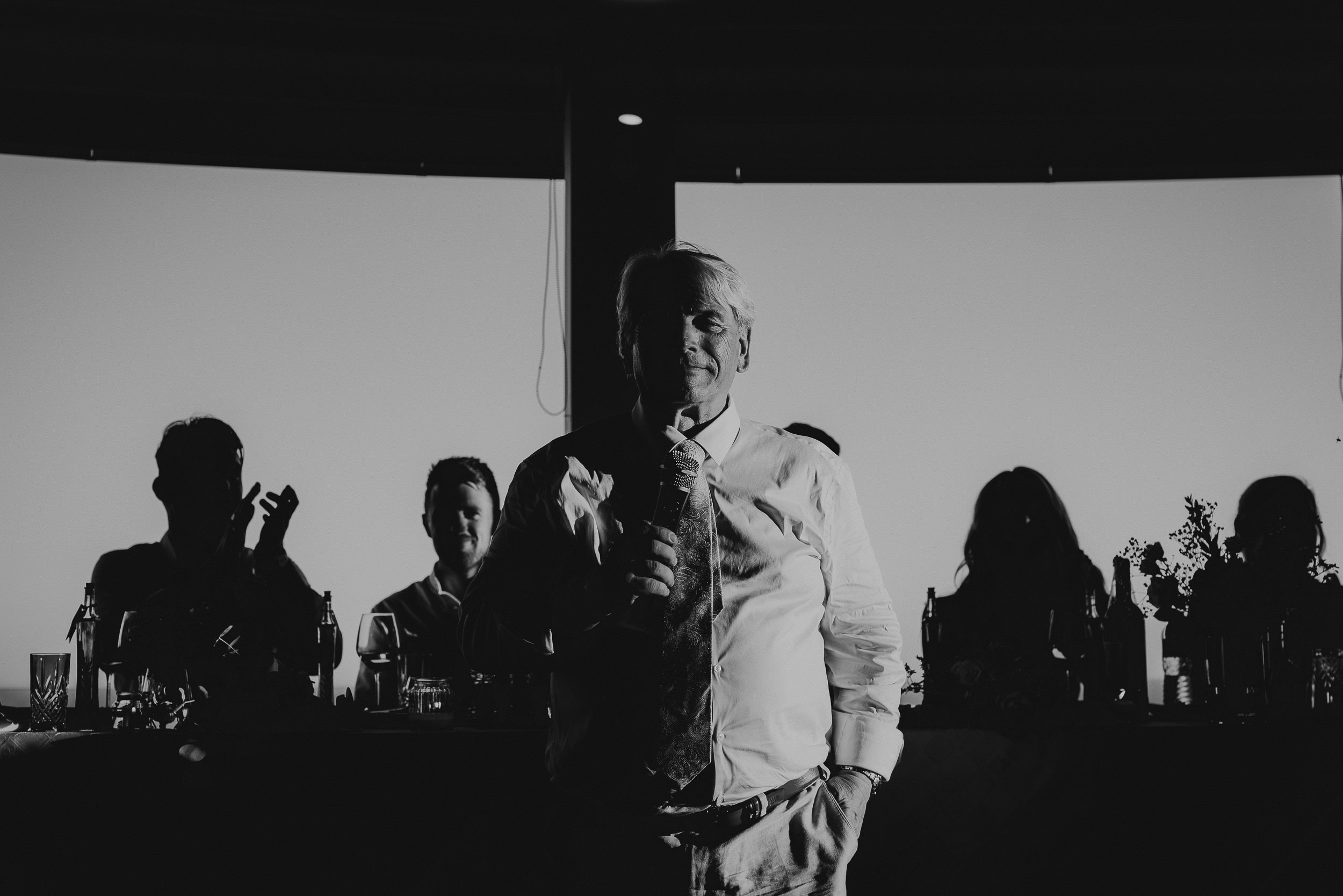A black and white wedding photo capturing a man standing in front of a group of people.