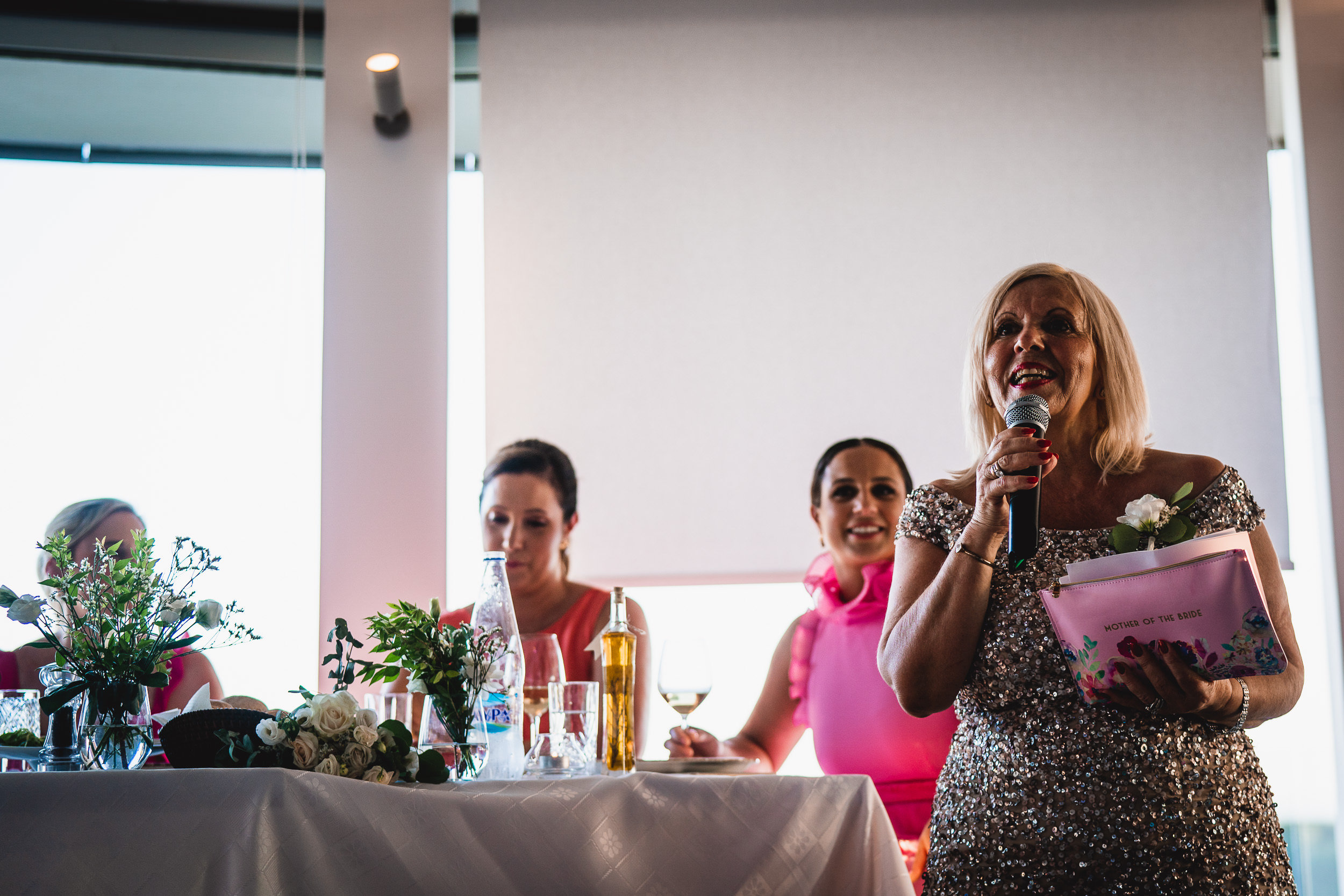 At a wedding celebration, a woman energetically addresses the crowd with a microphone as the groom and wedding photographer eagerly listen.