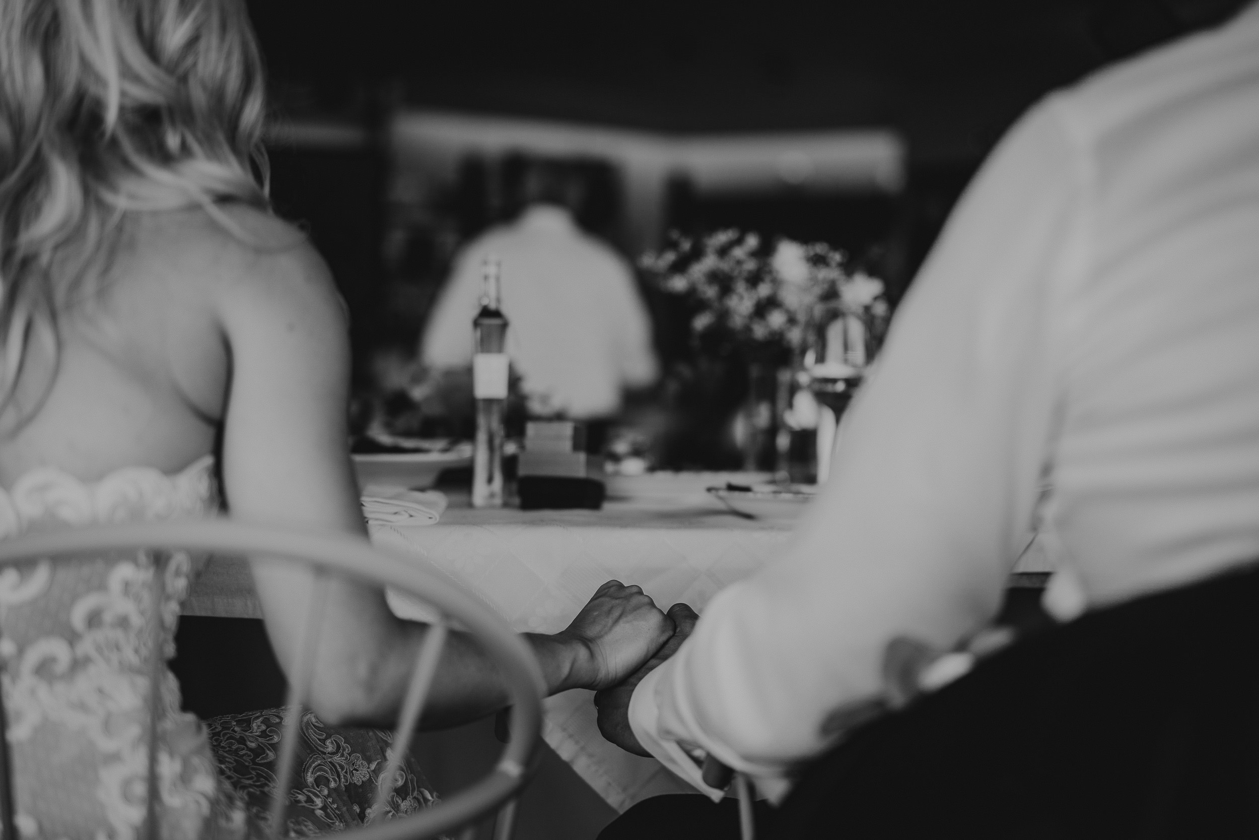 A wedding photographer captures the bride and groom's tender hand-holding moment at a table.