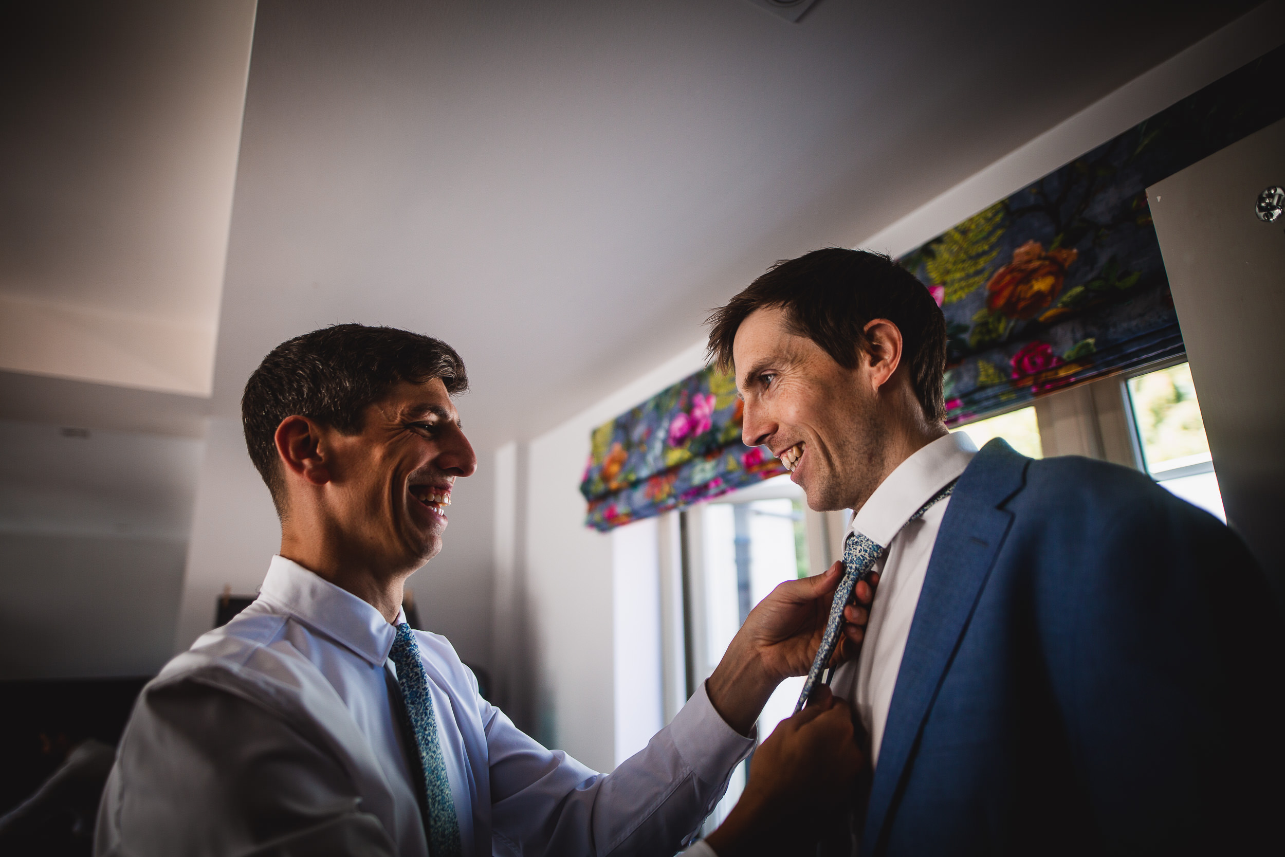 Two grooms getting ready for their wedding, tying their ties in front of a mirror while being photographed by a wedding photographer.