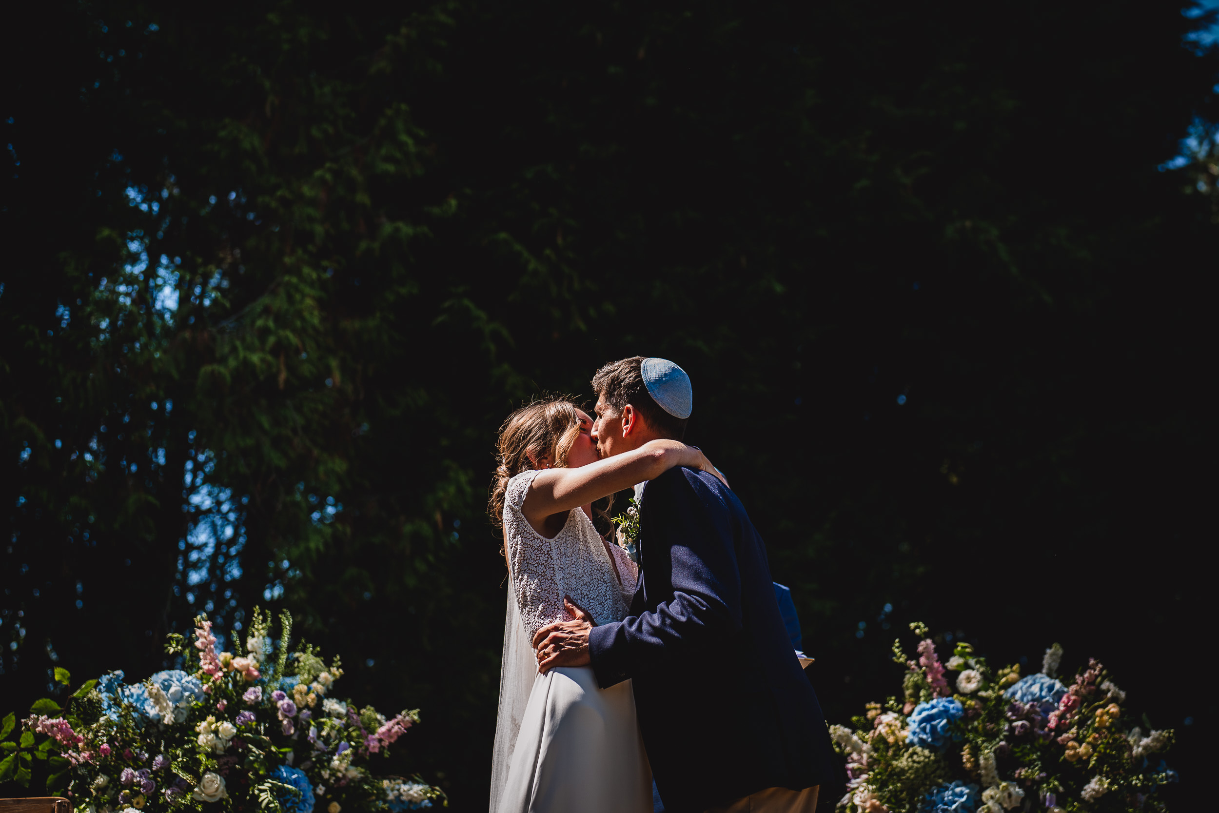 A wedding photographer capturing the groom and bride kissing under a tree in a garden.