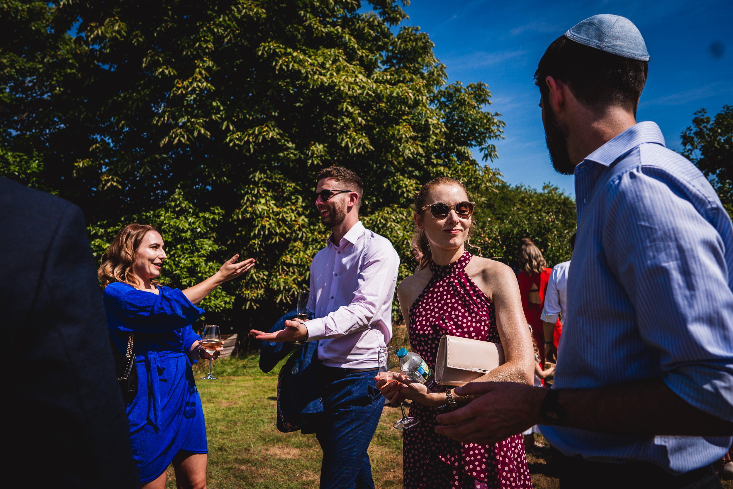 A wedding photographer captures a group of people talking to each other in a park during a wedding.