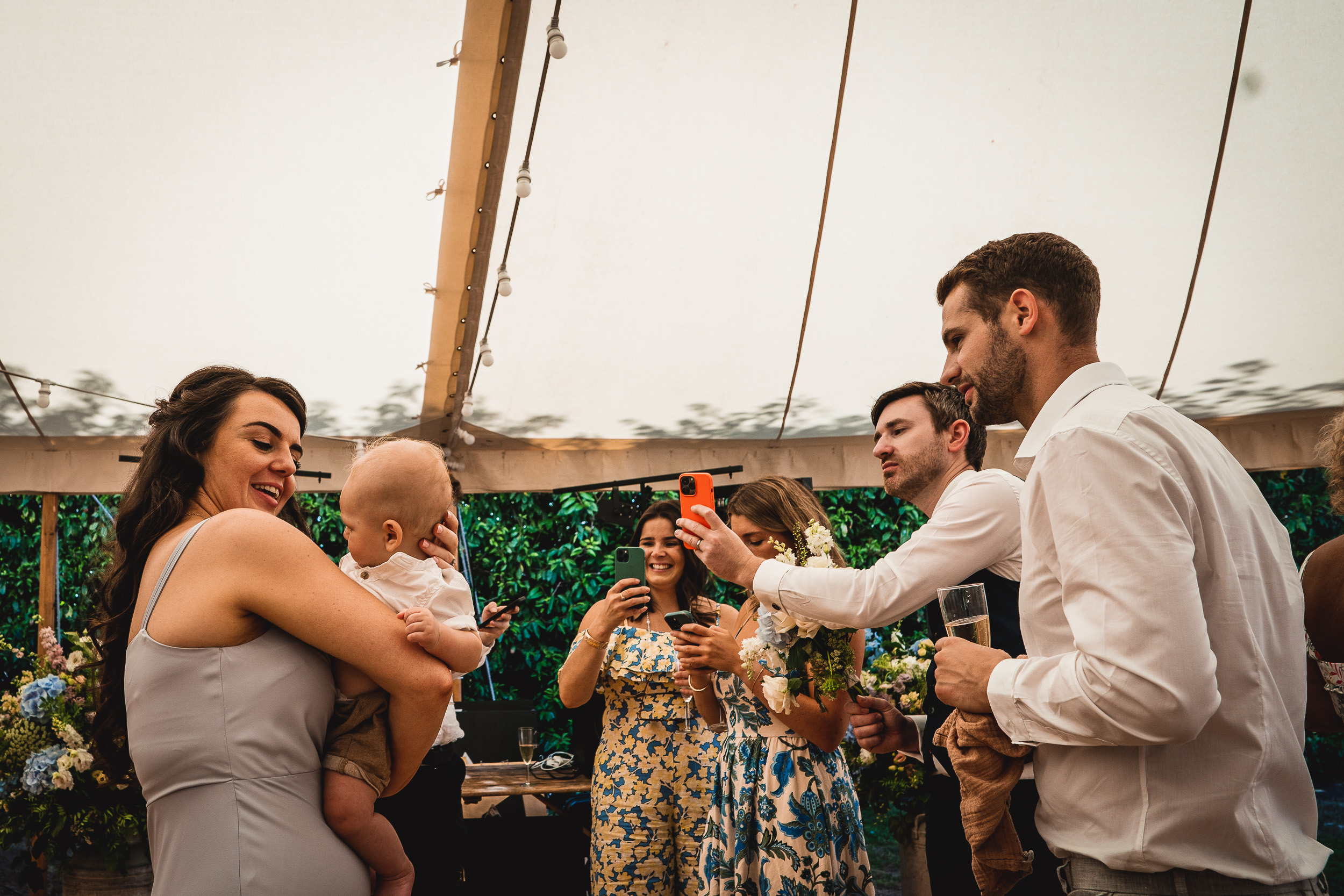 A bride, groom, and wedding photographer gather in a tent with a baby in their arms.
