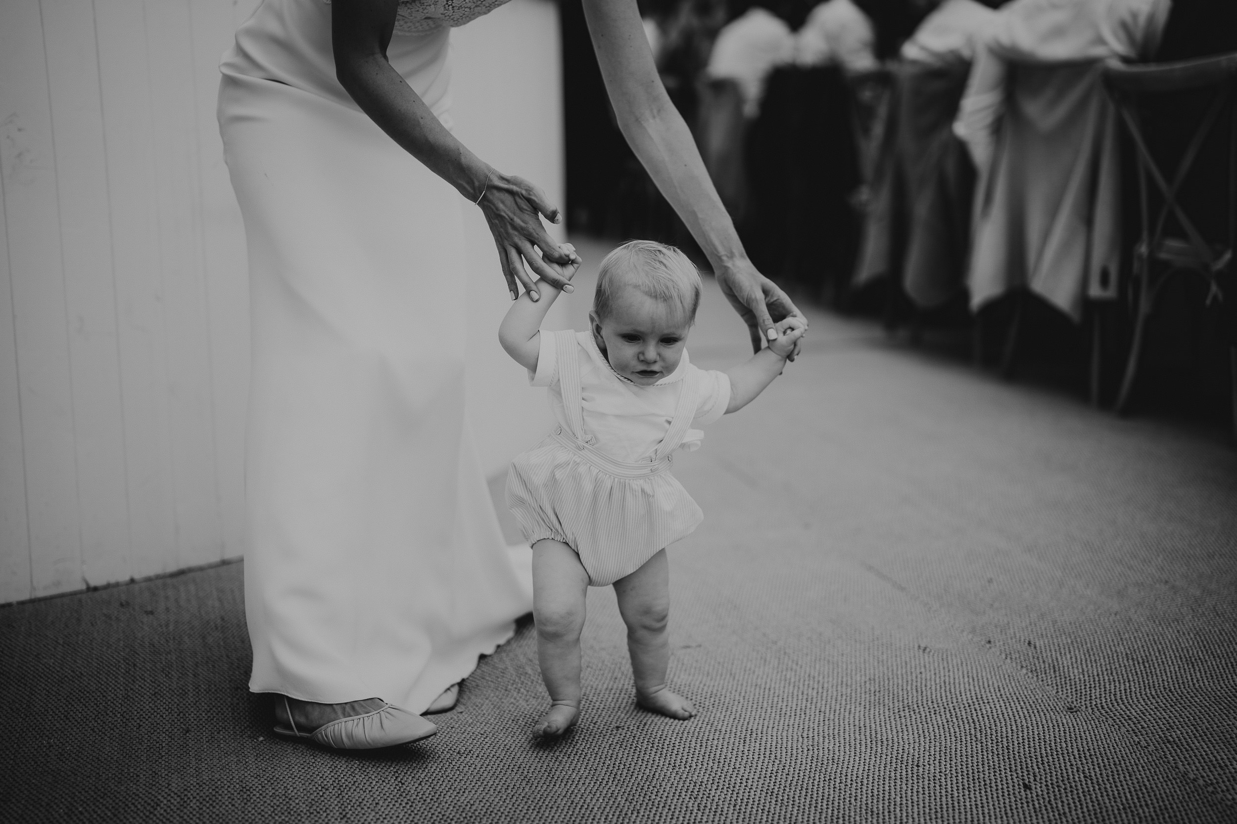 A woman holding a baby's hand walks down the aisle in a wedding photo.