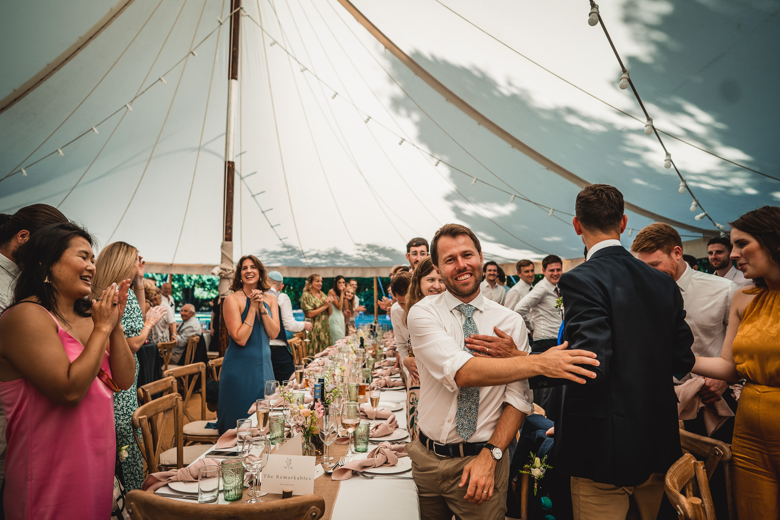 A wedding photographer captures joyful moments of the groom and guests at a reception in a tent.