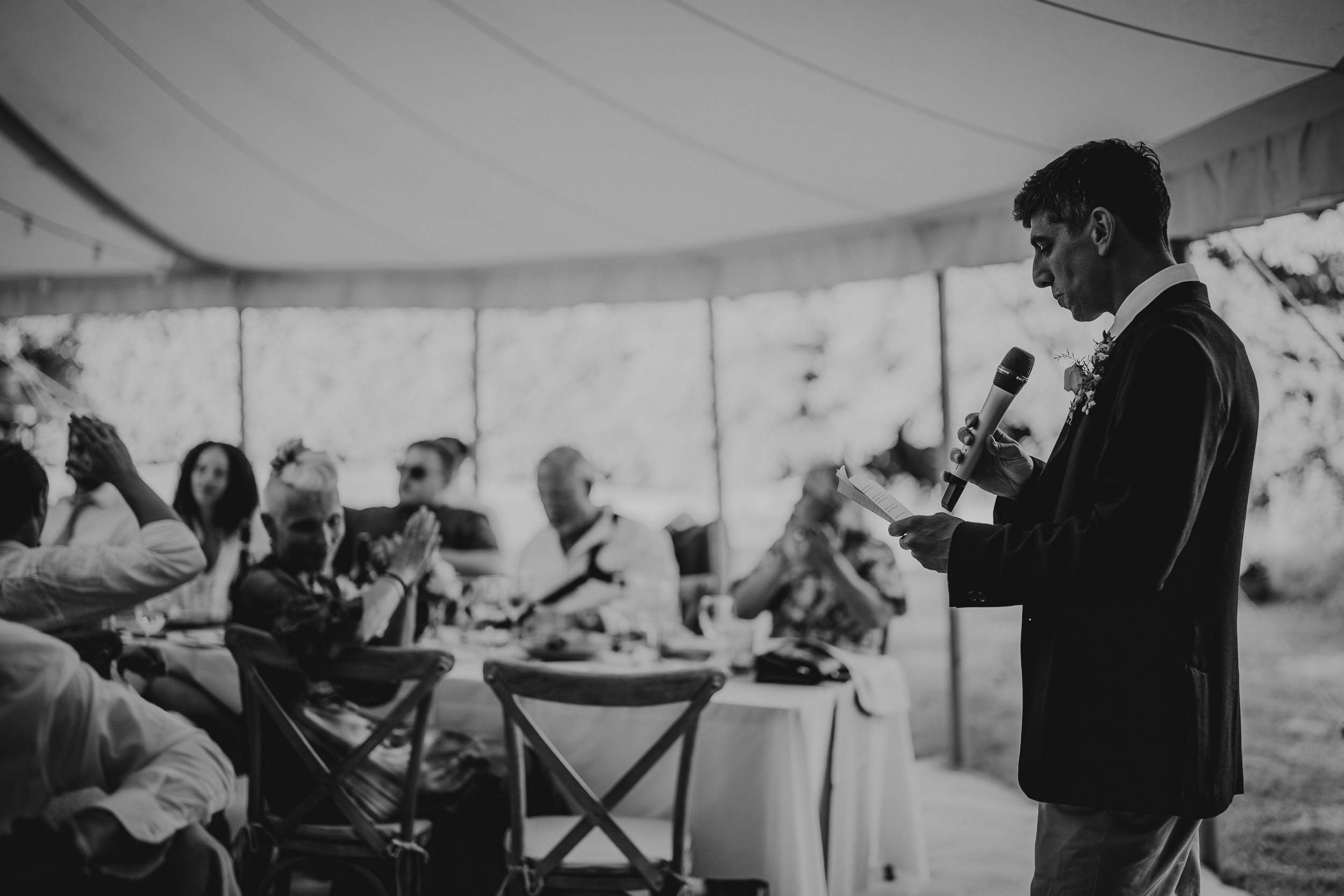 A wedding photographer captures a black and white photo of the groom while giving a speech at the wedding.